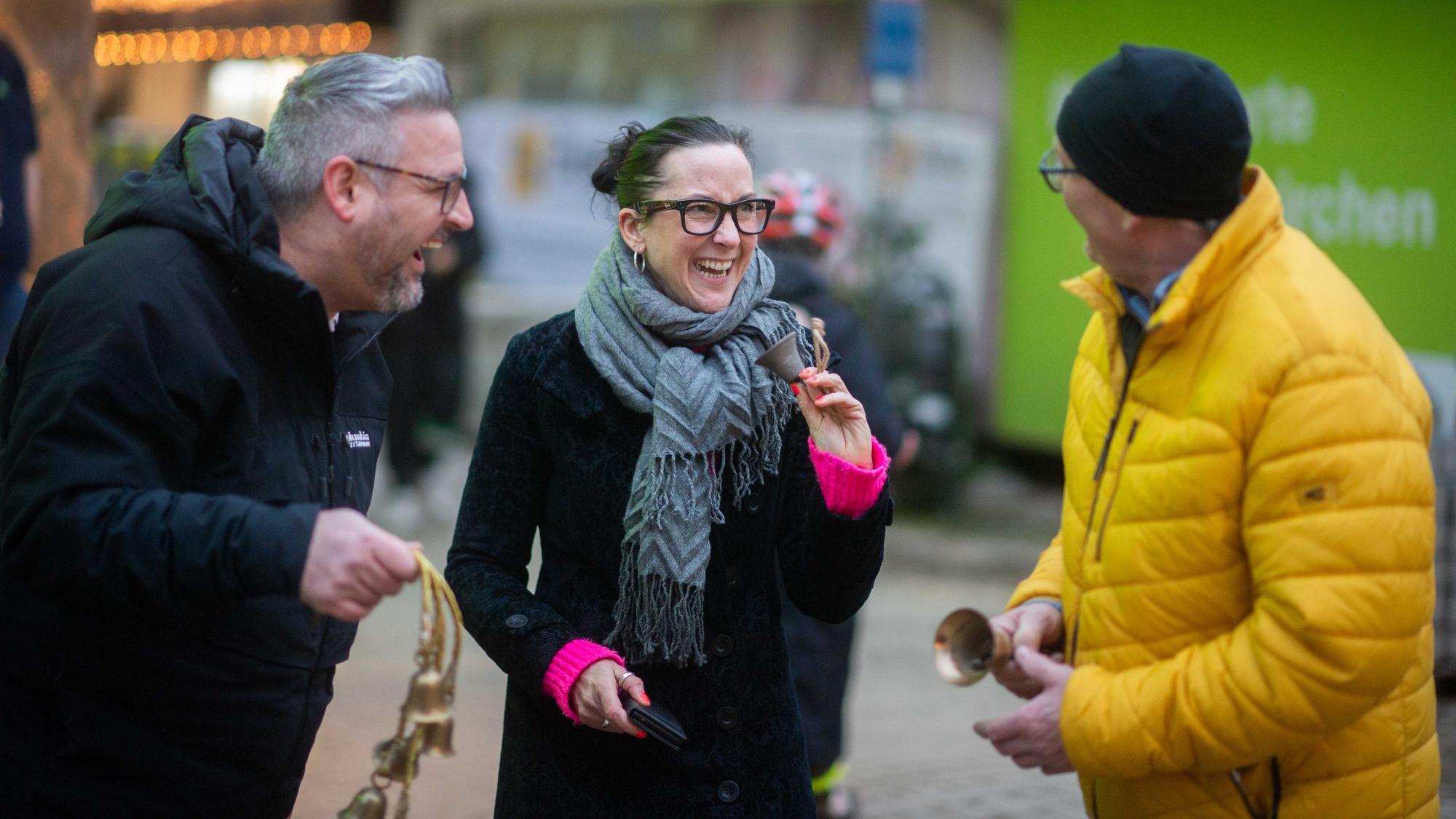 Das Bild zeigt die drei Initiatoren: Sie halten kleine Glocken in der Hand und lachen.