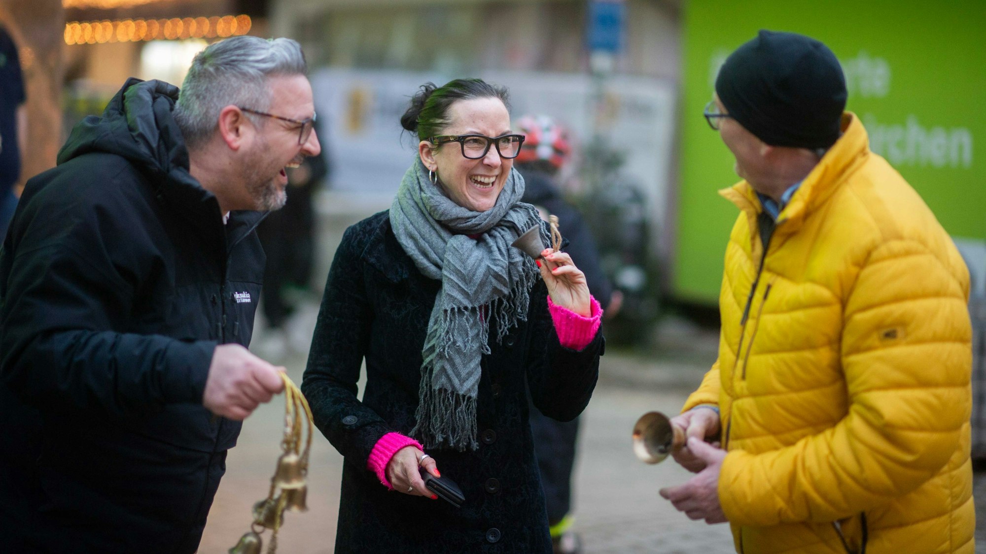Das Bild zeigt die drei Initiatoren: Sie halten kleine Glocken in der Hand und lachen.