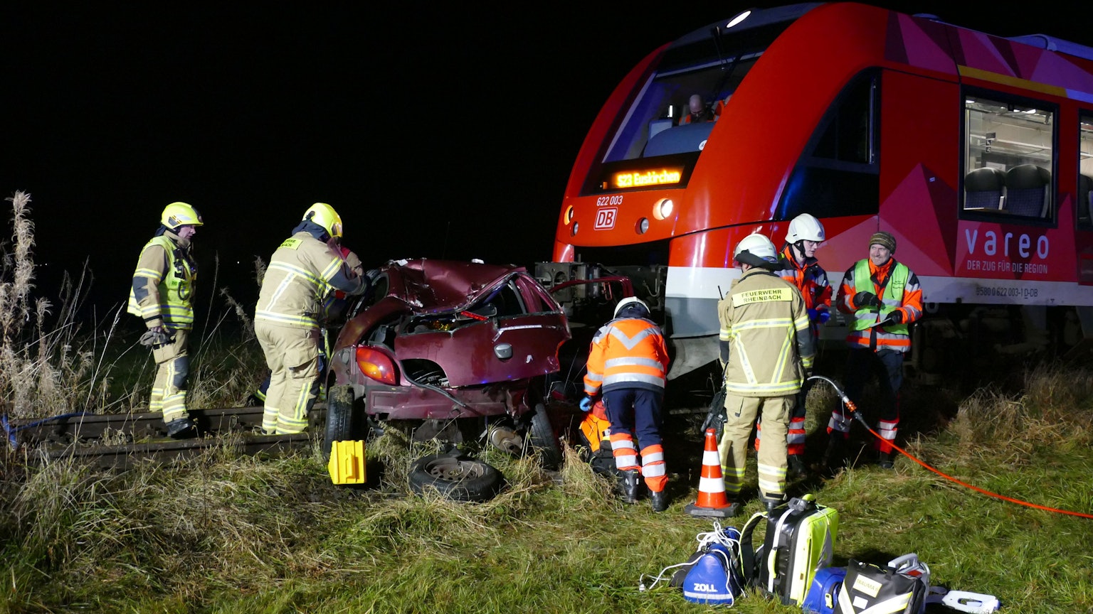 Feuerwehrleute stehen um ein zerstörtes Auto, das vor einem Zug steht.