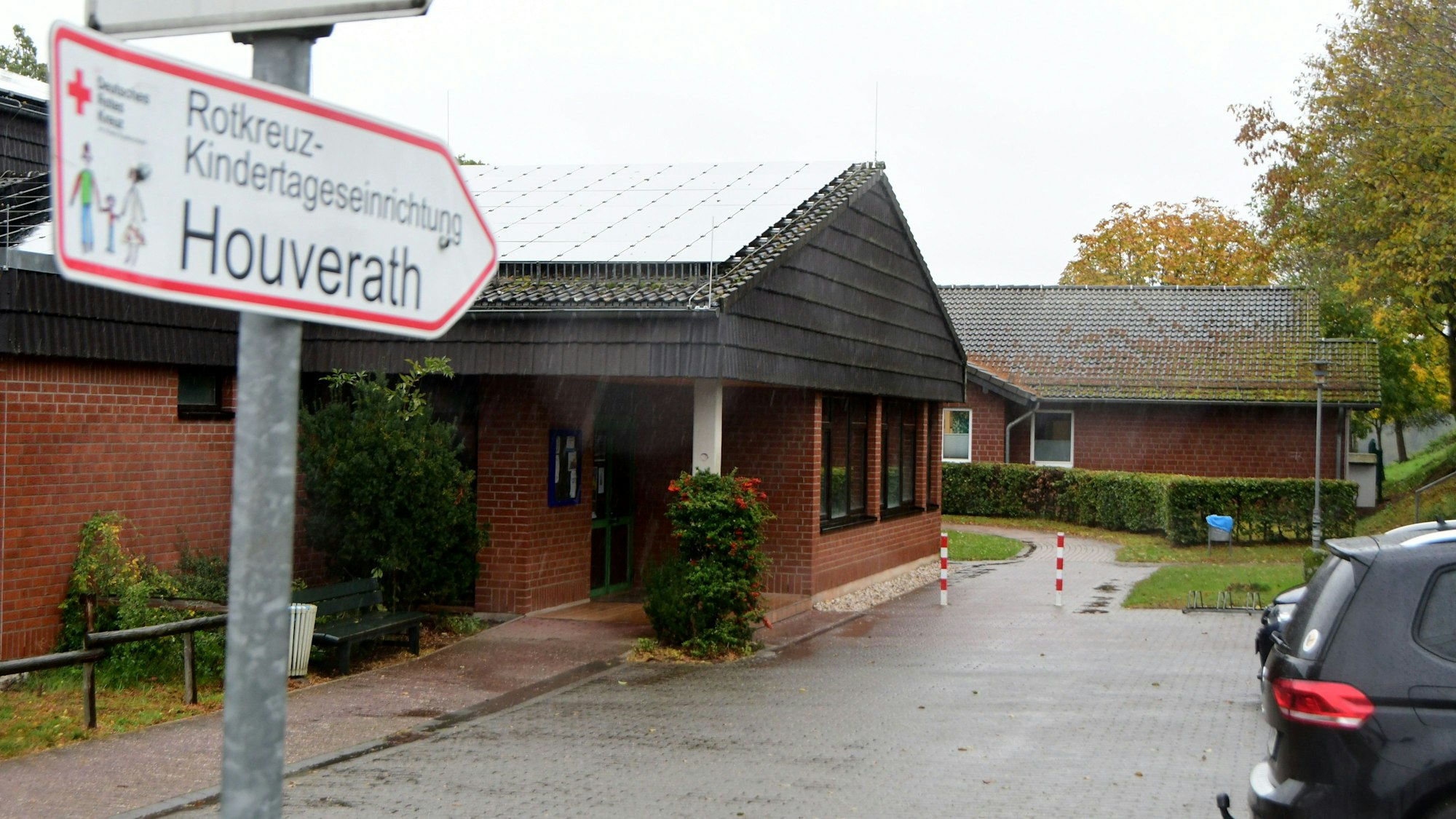 Auf einem Wegweiser steht "Rotkreuz-Kindertageseinrichtung Houverath", dahinter ist der Kindergarten zu sehen.