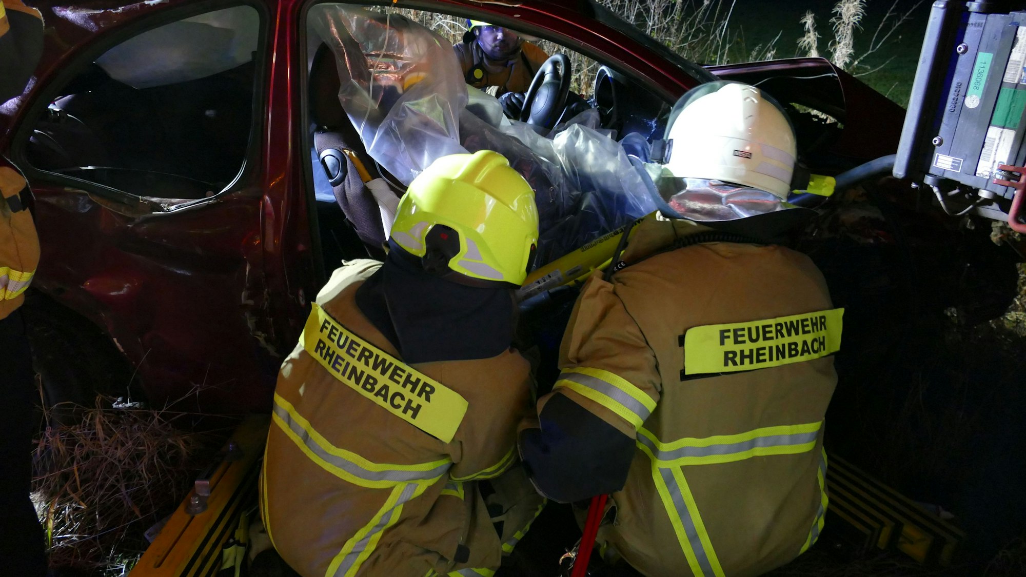 Feuerwehrleute hocken vor einem Autowrack, in dem ein Verletzter sitzt, der mit einer Plane vor Splittern geschützt wird.
