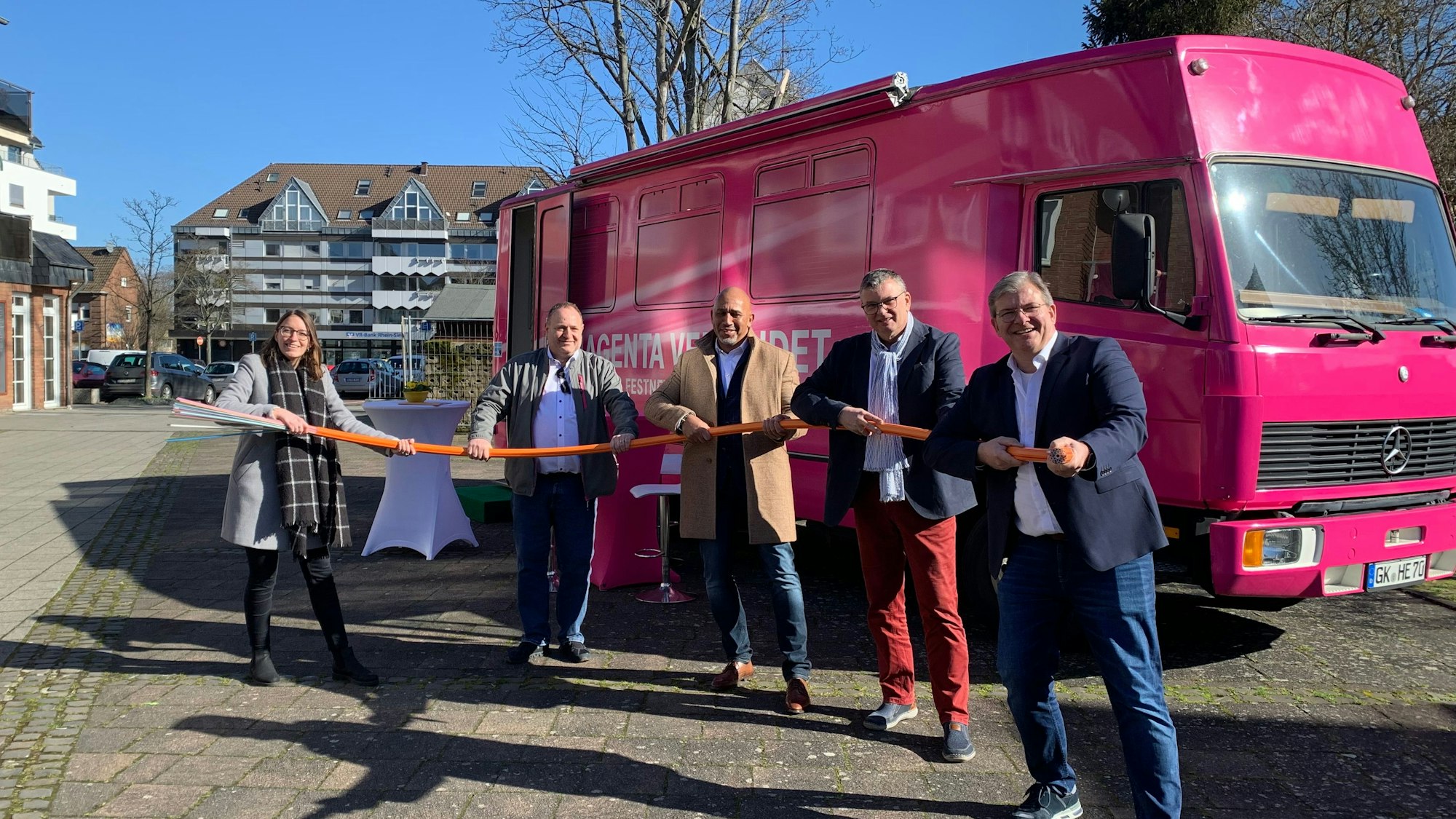 Bei einem Pressetermin für den Glasfaserausbau stehen Vertreter der Stadt und der Telekom vor einem magentafarbenen Bus und halten ein meterlanges Glasfaserkabel in der Hand.