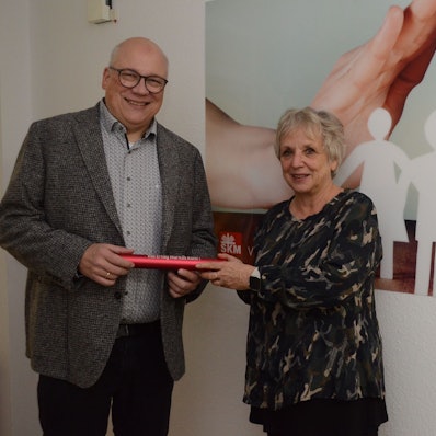 Ein Mann mit Glatze und Brille nimmt einen roten Staffelstab aus Metall in Empfang. Eine Frau mit grauen Haaren überreicht das Rohr.
