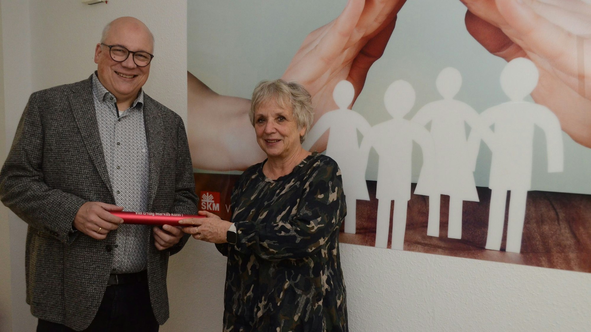 Ein Mann mit Glatze und Brille nimmt einen roten Staffelstab aus Metall in Empfang. Eine Frau mit grauen Haaren überreicht das Rohr.