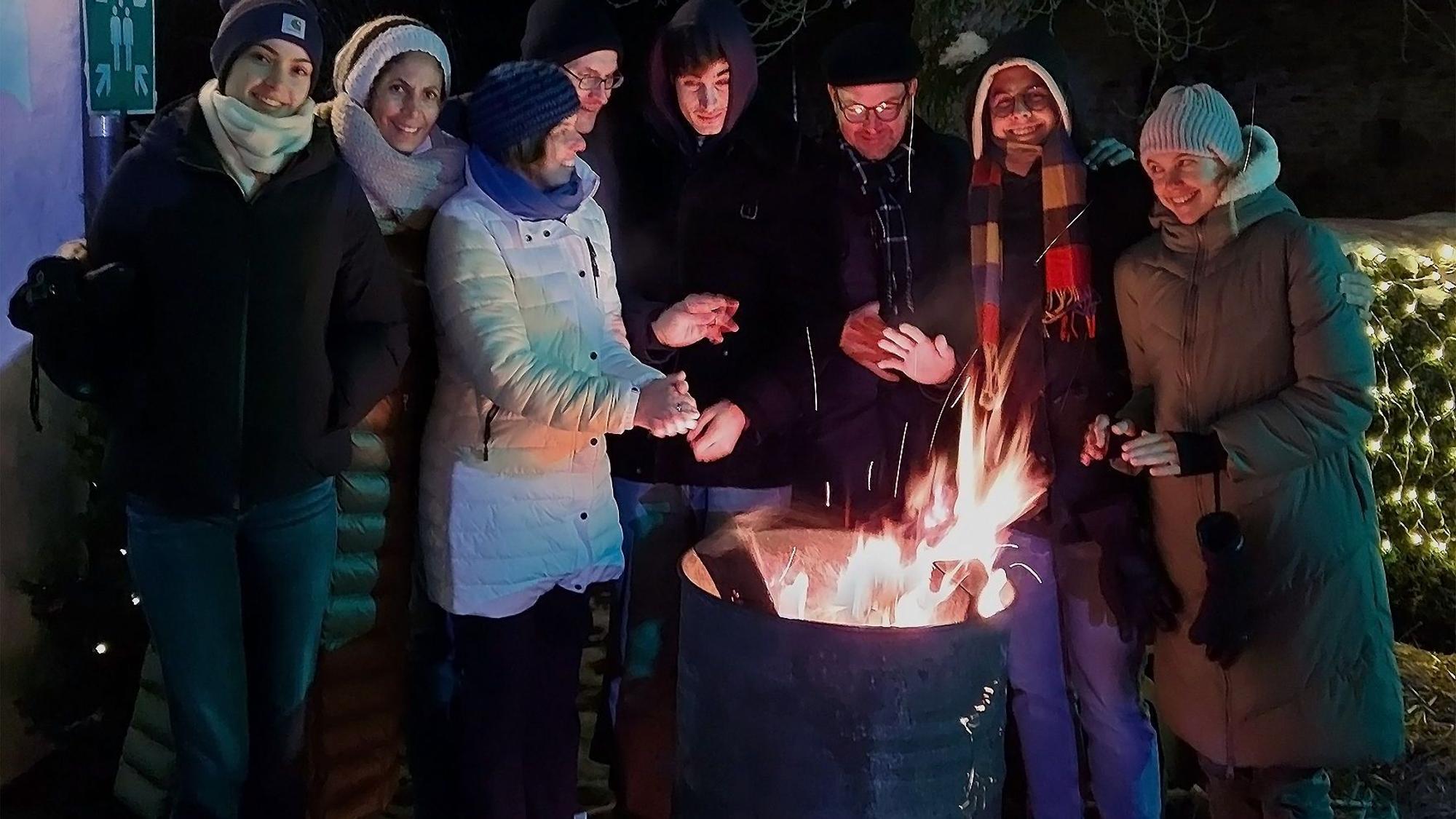 Acht Leute stehen an einem Feuer und wärmen sich.