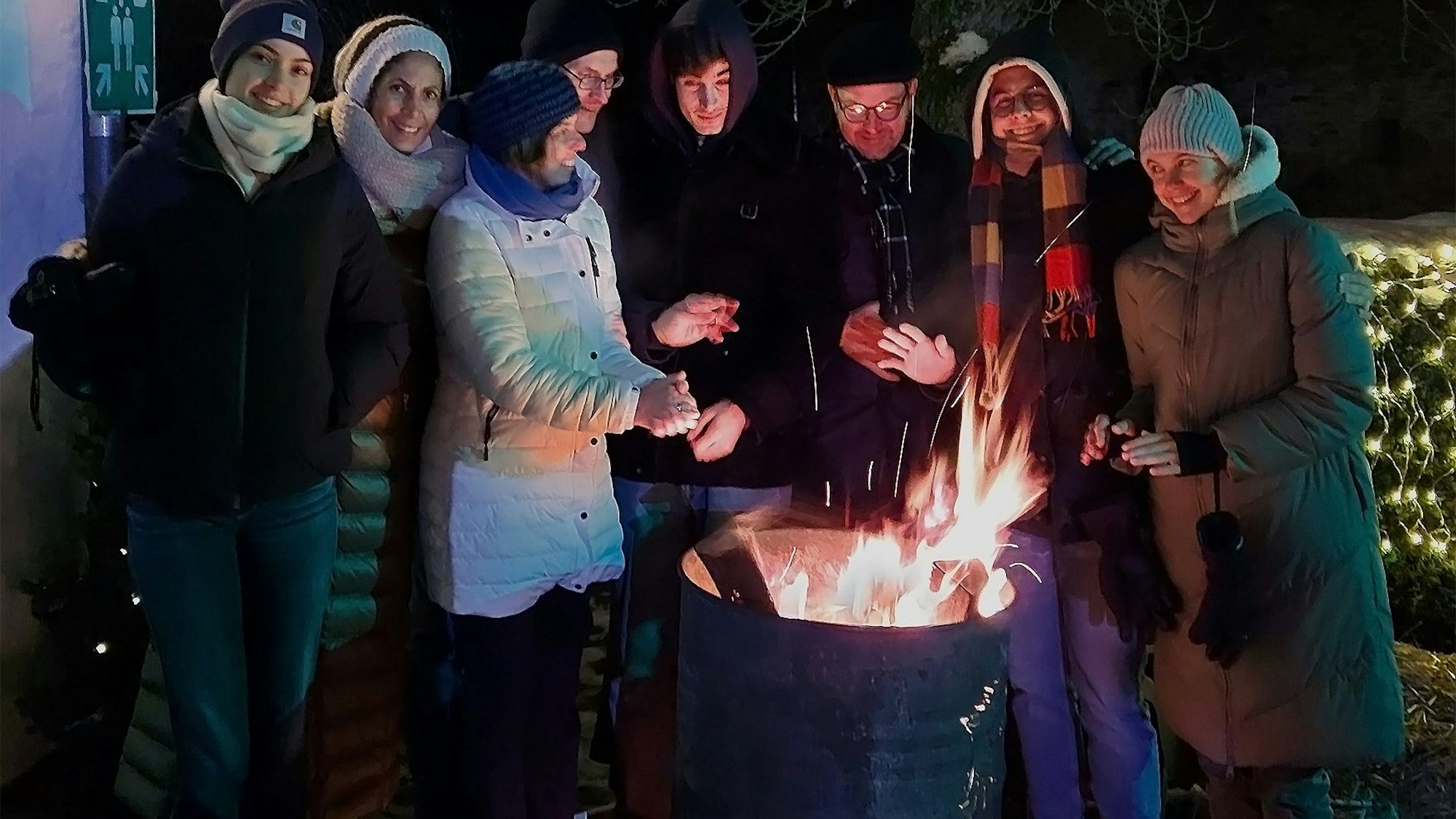 Acht Leute stehen an einem Feuer und wärmen sich.