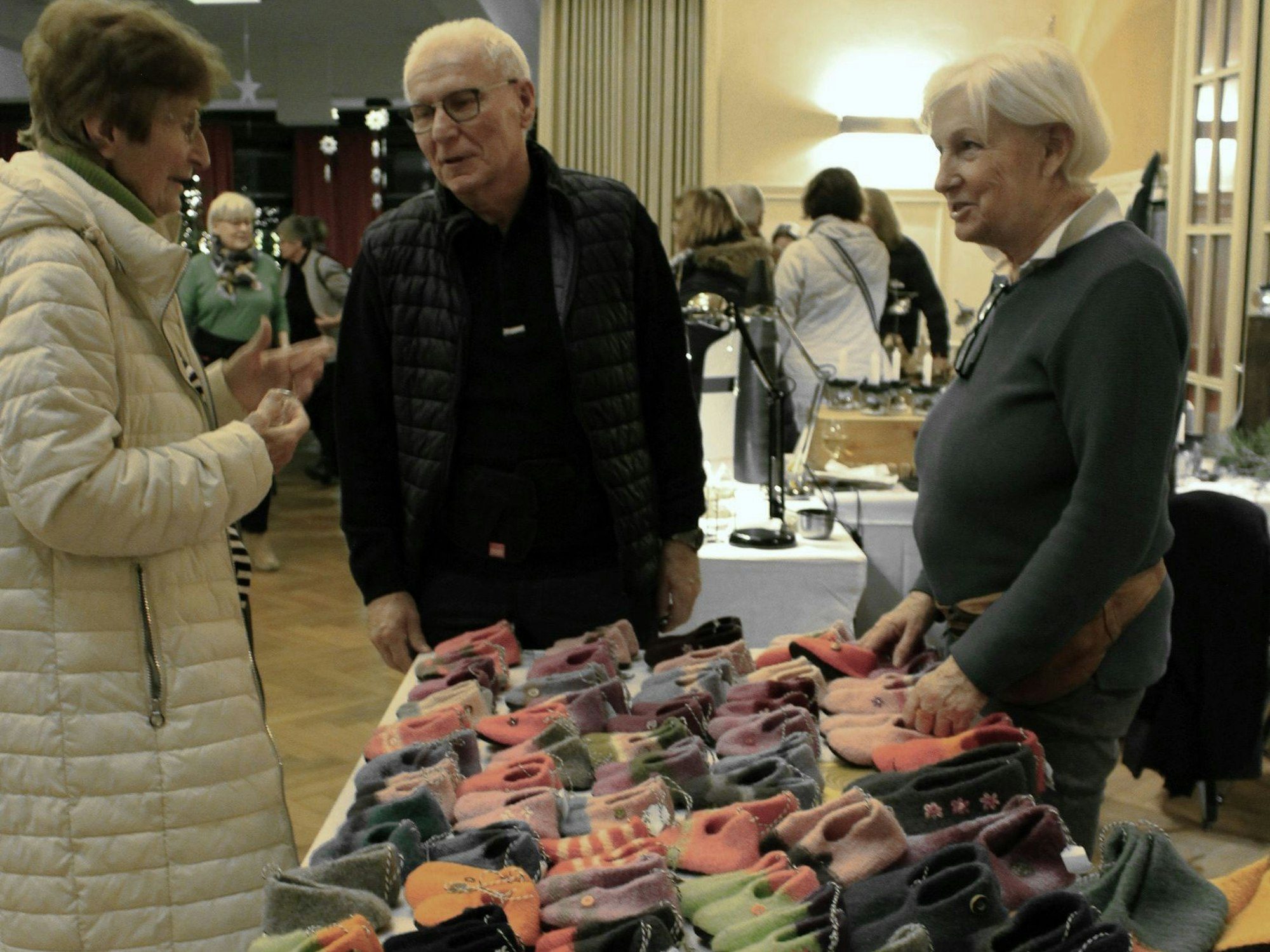 Das Bild zeigt Meggie und Ernst Flacke an ihrem Stand. Auf dem Tisch liegen unzählige Hausschuhe. Eine Besucherin lässt sich beraten.