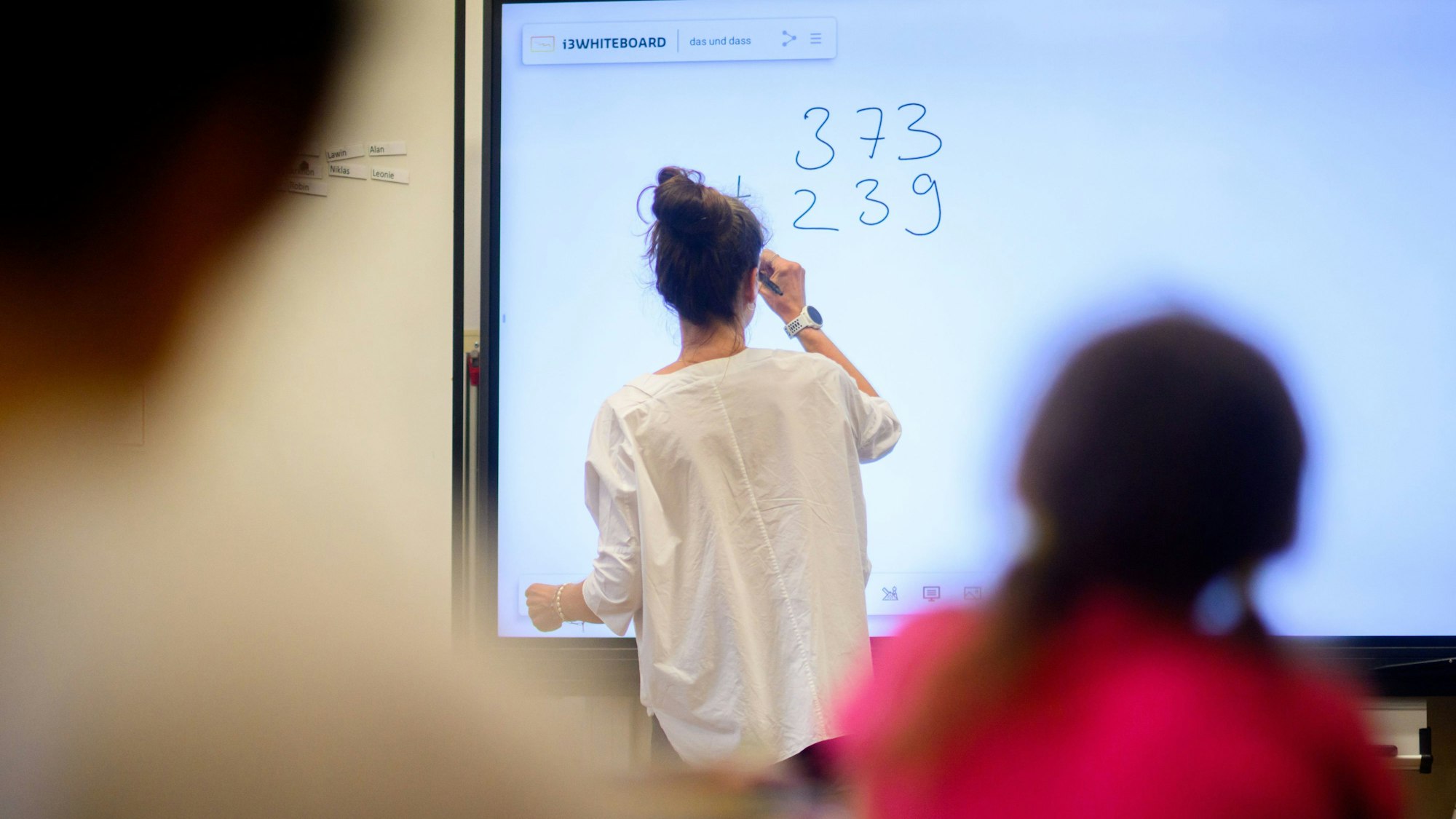 Eine Lehrerin schreibt eine Mathematikaufgabe auf eine digitale Schultafel im Klassenraum einer 4. Klasse einer Grundschule.