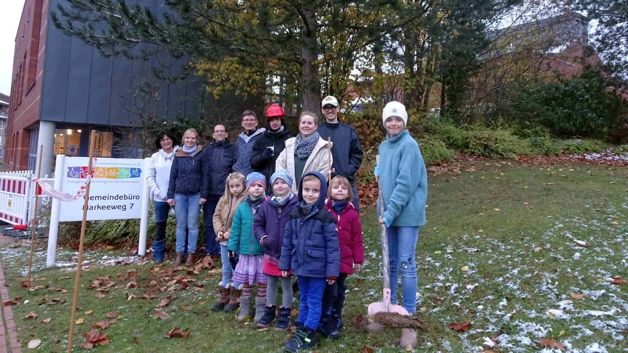 Das inklusive Team der evangelischen Kirche Meckenheim um Diakonin Sonja Freischem (helle jacke Mitte), kleine Sängerinnen und Sänger aus dem Kinderchor, Mitglieder und Betreuer der inklusiven Gruppen, Pfarrer Stefan Bergner (4.v.l.), Baukirchmeister Michael Blum (links daneben) sowie Dr. Simone Gangl (2.v.l.) als Vorsitzende des Presbyteriums stehen vor dem Gemeindezentrum in Meckenheim und stechen mit einem Spaaten in die Erde.