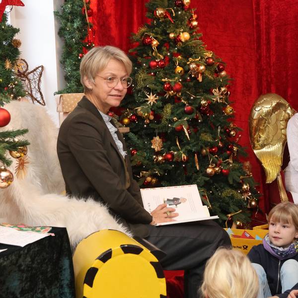 NRW-Schulministerin Dorothee Feller sitzt mit einem aufgeschlagenen Buch auf dem Schoß vor einem geschmückten Weihnachtsbaum. Kinder und das Christkind hören ihr zu.