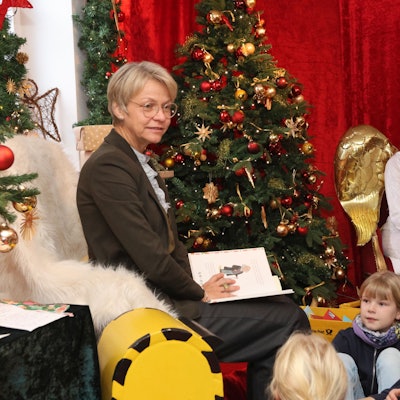 NRW-Schulministerin Dorothee Feller sitzt mit einem aufgeschlagenen Buch auf dem Schoß vor einem geschmückten Weihnachtsbaum. Kinder und das Christkind hören ihr zu.
