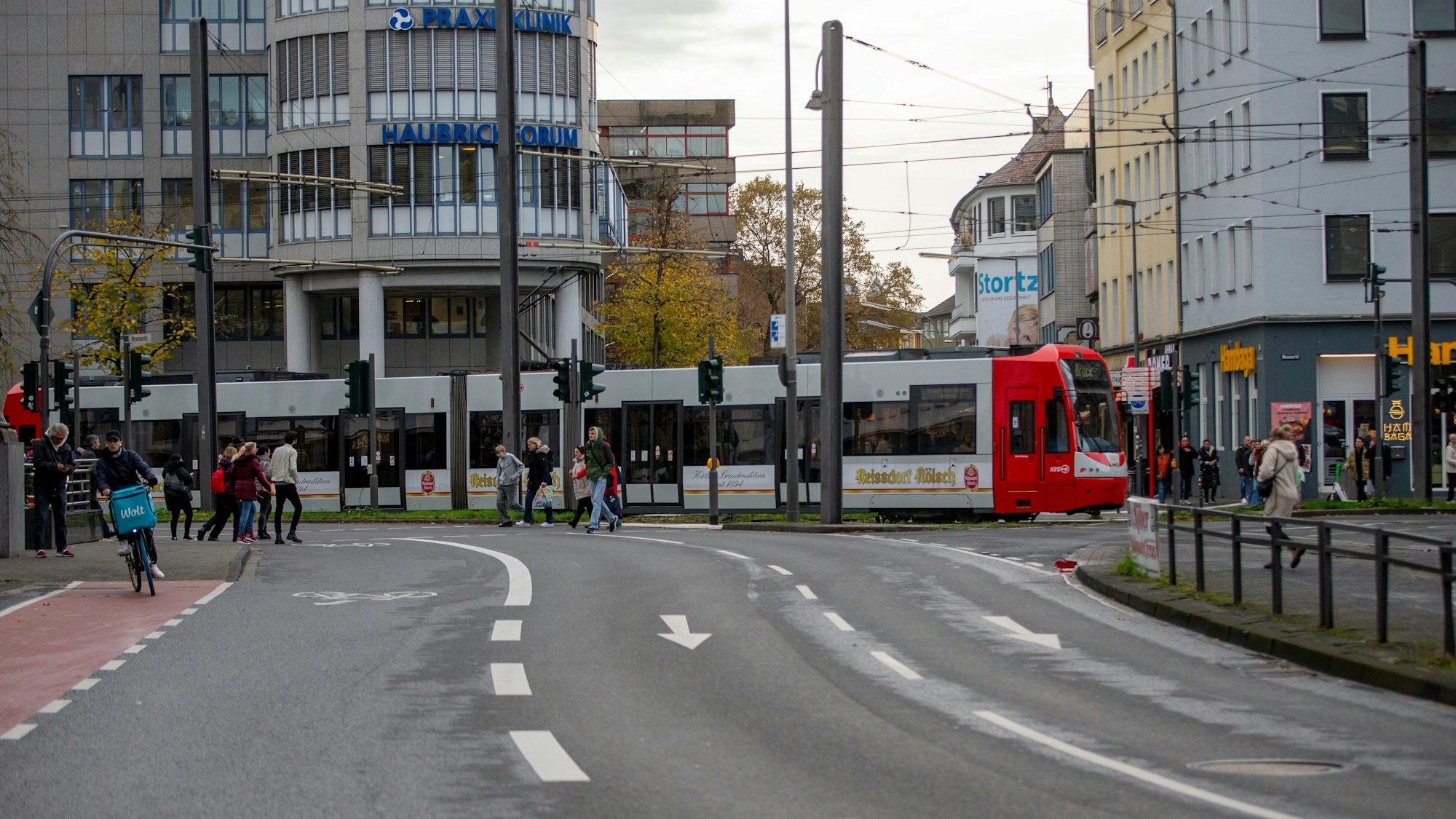 Eine Straßenbahn fährt eine leichte Kurve auf den Neumarkt zu. Fußgänger gehen über eine Straße.