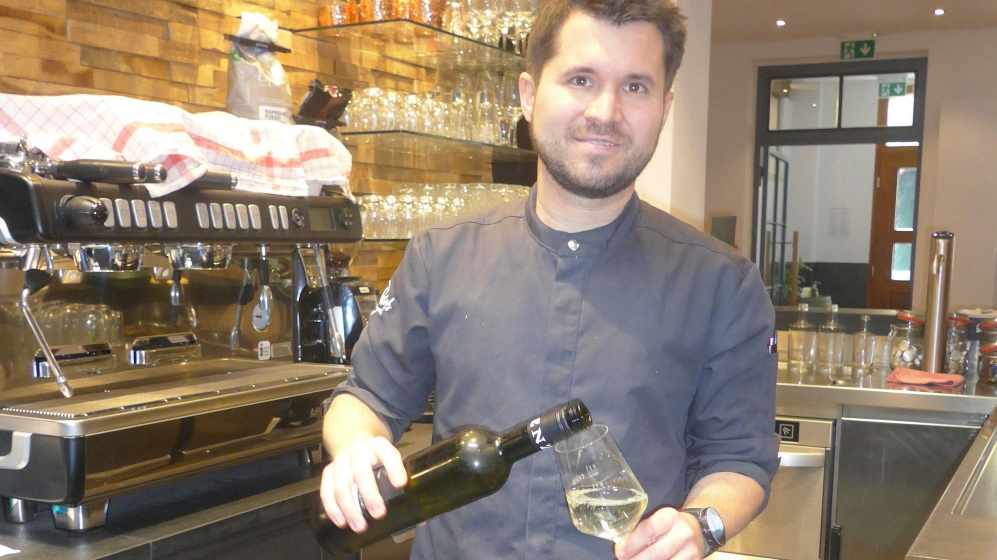 Auf dem Foto ist der Bedburger Gastronom Christof Hölscher zu sehen. Er schenkt Weißwein aus einer Flasche in ein Glas ein.