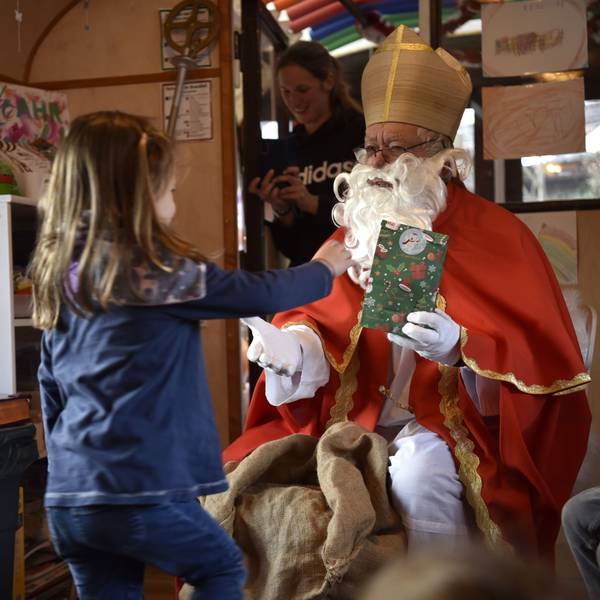 Ein Mann mit rotem Mantel, Mitra auf dem Kopf und langem, weißen Bart hält einem Mädchen eine bunte Geschenk-Tüte hin.