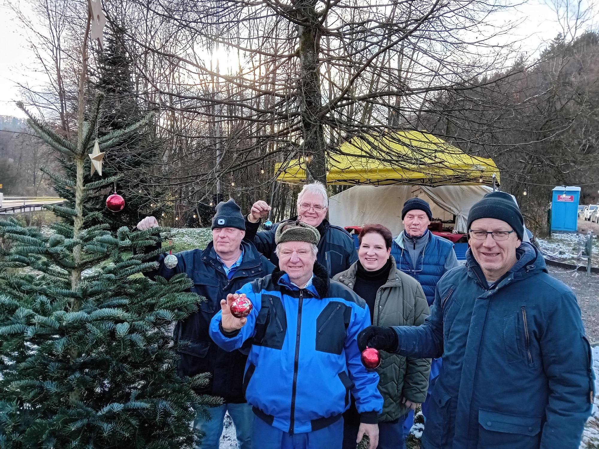 Männer in Arbeitskleidung stehen neben einem Tannenbaum und zeigen Christbaumschmuck.
