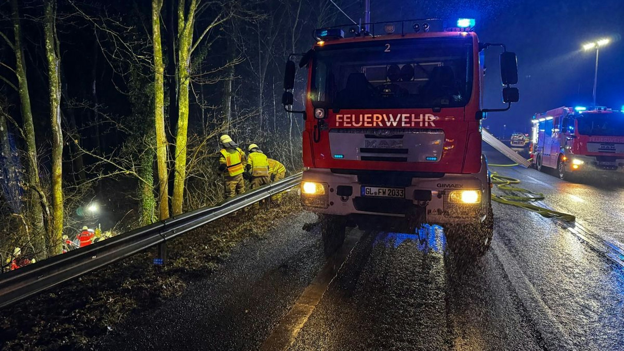 Feuerwehrautos stehen auf der Overather Straße, Feuerwehrleute arbeiten in der Böschung neben der Straße.