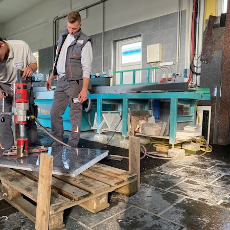 In einer Werkstatt bearbeitet der Azubi Pascal Elschenbroich eine polierte Natursteinplatte mit einem großen Bohrer. Seine Arbeit wird von einem Kollegen beobachtet.
