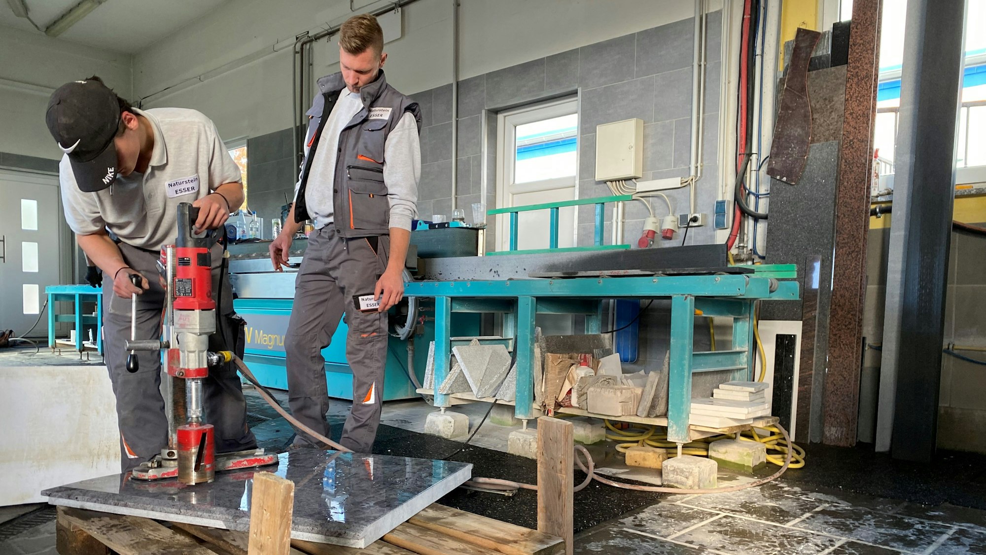 In einer Werkstatt bearbeitet der Azubi Pascal Elschenbroich eine polierte Natursteinplatte mit einem großen Bohrer. Seine Arbeit wird von einem Kollegen beobachtet.