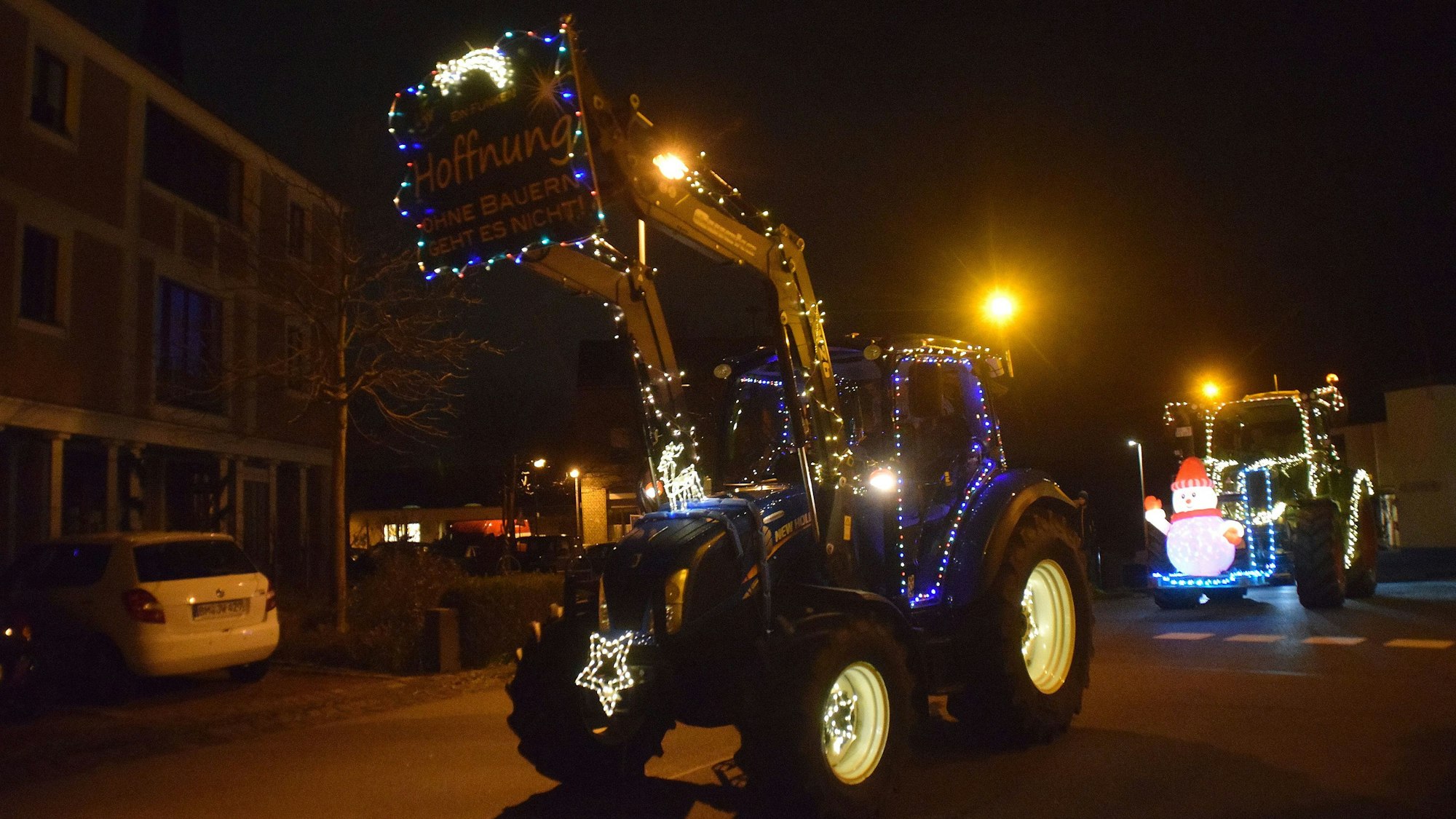 Mehrere mit Lichterketten und bunten Figuren geschmückte Traktoren fahren bei Dunkelheit durch eine Straße.