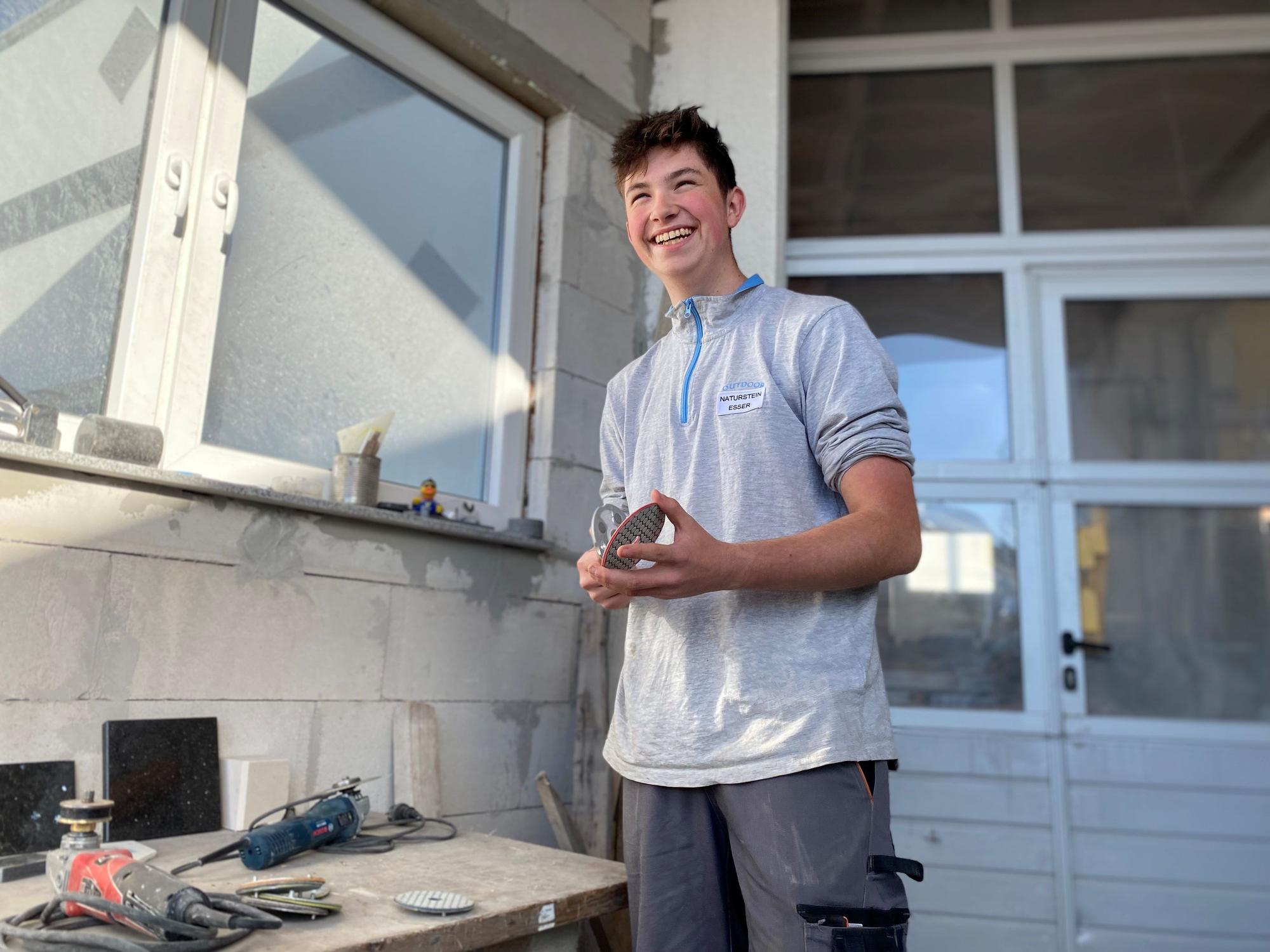 Naturwerksteinmechanik-Azubi Benjamin Achnitz hält ein Schleifpapier in den Händen und lacht über den Scherz eines Kollegen.