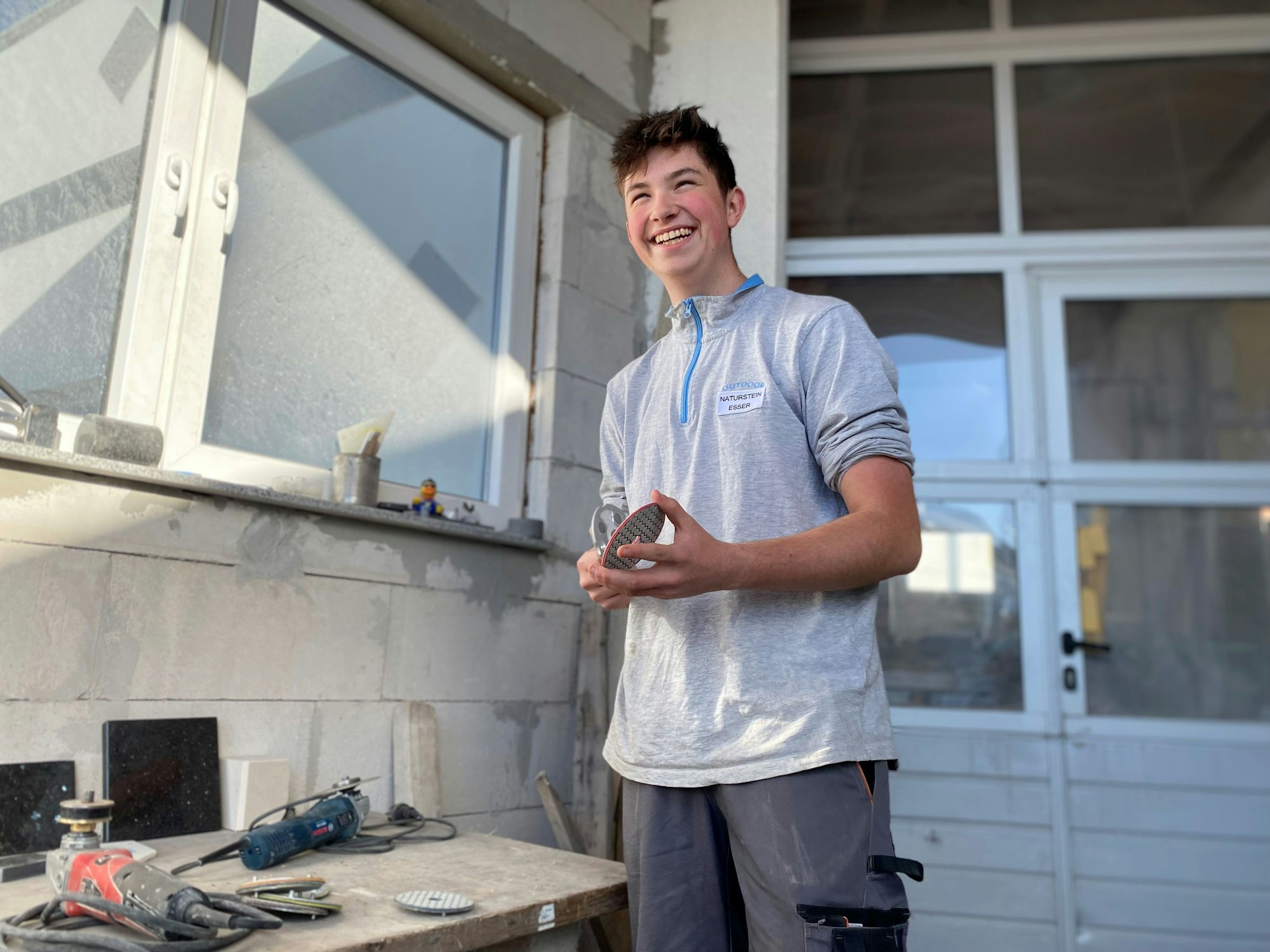 Naturwerksteinmechanik-Azubi Benjamin Achnitz hält ein Schleifpapier in den Händen und lacht über den Scherz eines Kollegen.