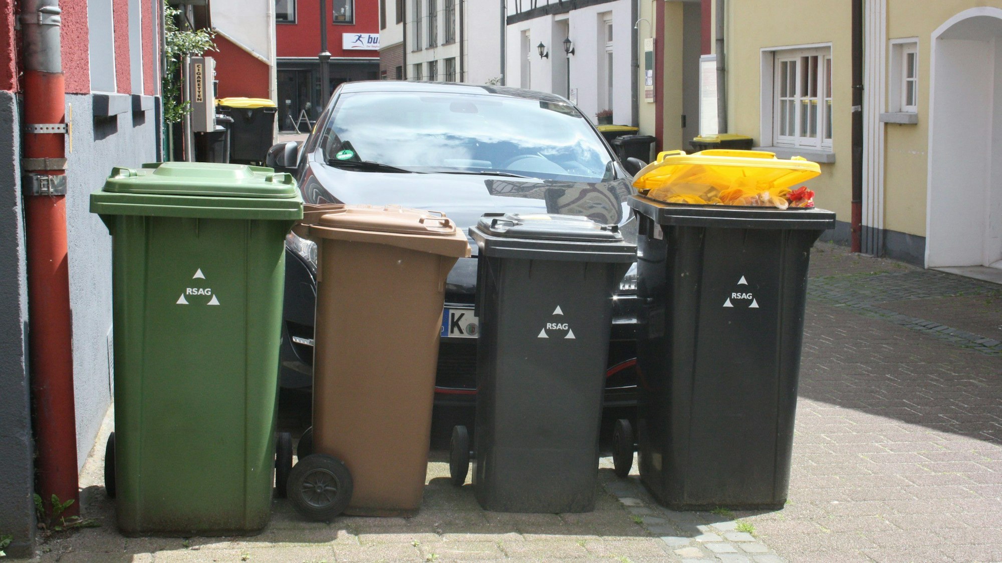 Eine Papiertonne, eine Biotonne, eine Restmülltonne und eine Wertstofftonne der RSAG stehen nebeneinander abholbereit auf einer Straße in Siegburg.