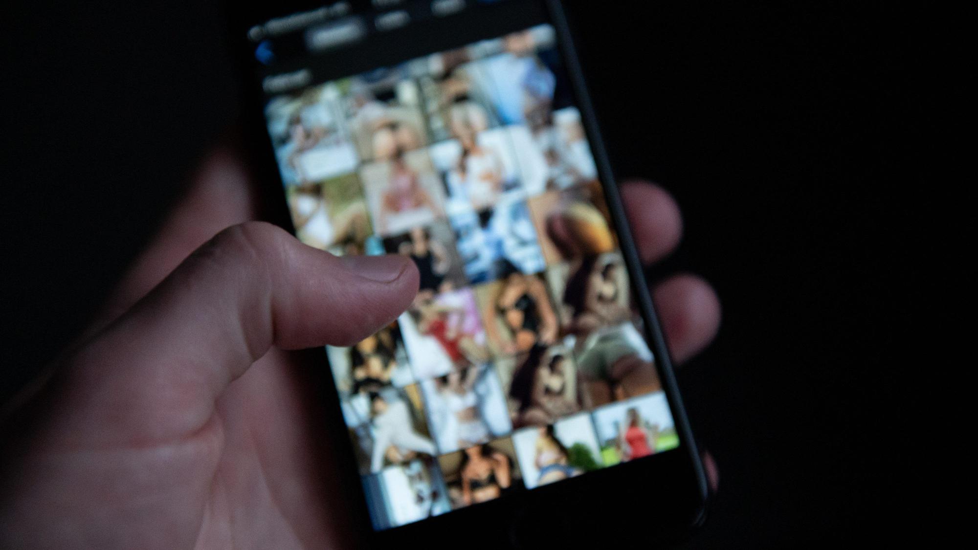 Eine Hand hält ein Handy, der Bildschirm zeigt pornografische Fotos.