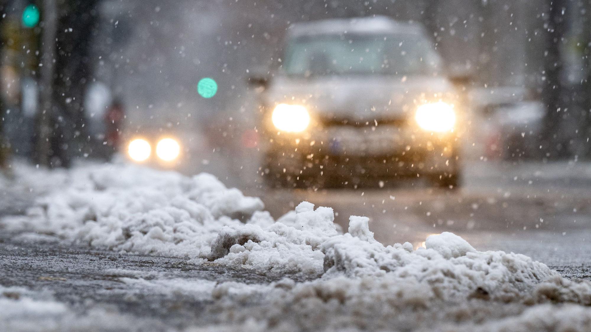 Schnee und Eis liegen am Straßenrand, während ein Auto durch den Schnee fährt. Der Deutsche Wetterdienst (DWD) erwartet zum Wochenende Neuschnee und Eisregen in Köln und der Region. Autofahrer könnten durch den gefrierenden Regen überrascht werden. (Symbolbild)