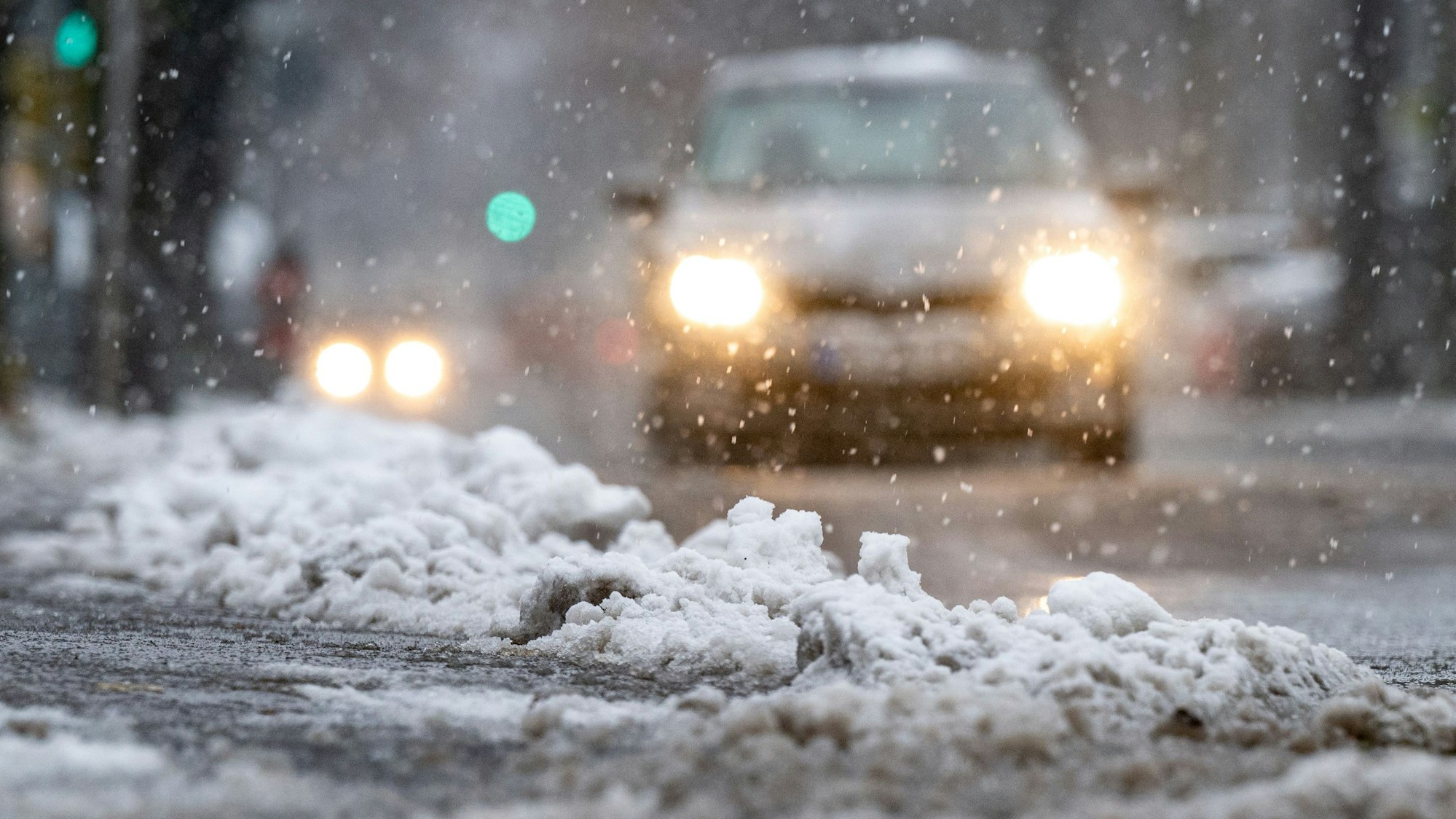 Schnee und Eis liegen am Straßenrand, während ein Auto durch den Schnee fährt. Der Deutsche Wetterdienst (DWD) erwartet zum Wochenende Neuschnee und Eisregen in Köln und der Region. Autofahrer könnten durch den gefrierenden Regen überrascht werden. (Symbolbild)