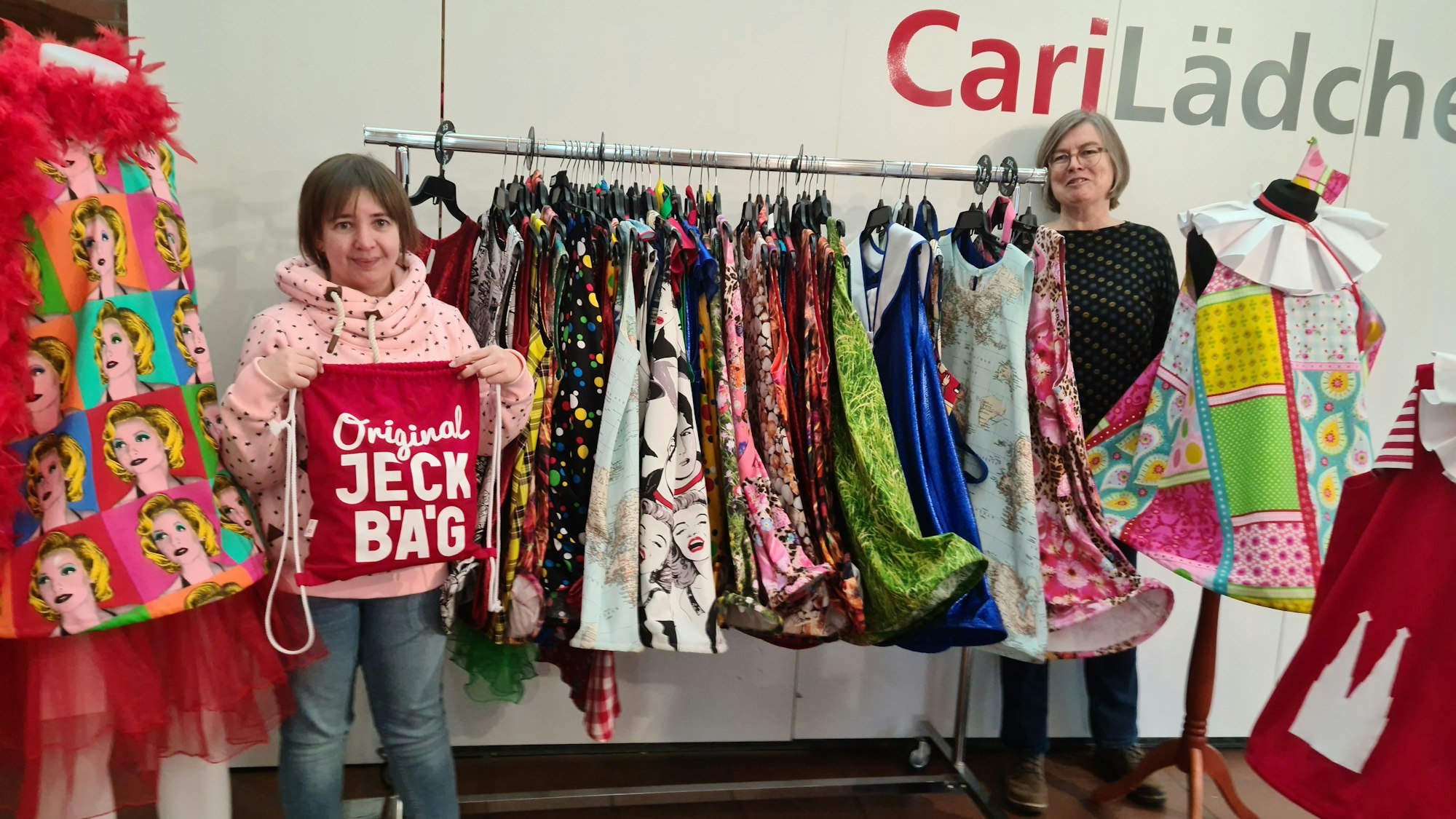 Eine junge Frau hält eine Stofftasche mit dem Schriftzug Original Jeck Bäg, eine ältere Frau steht neben einer Kleiderstange mit vielen Kleidern in verschiedenen Farben.