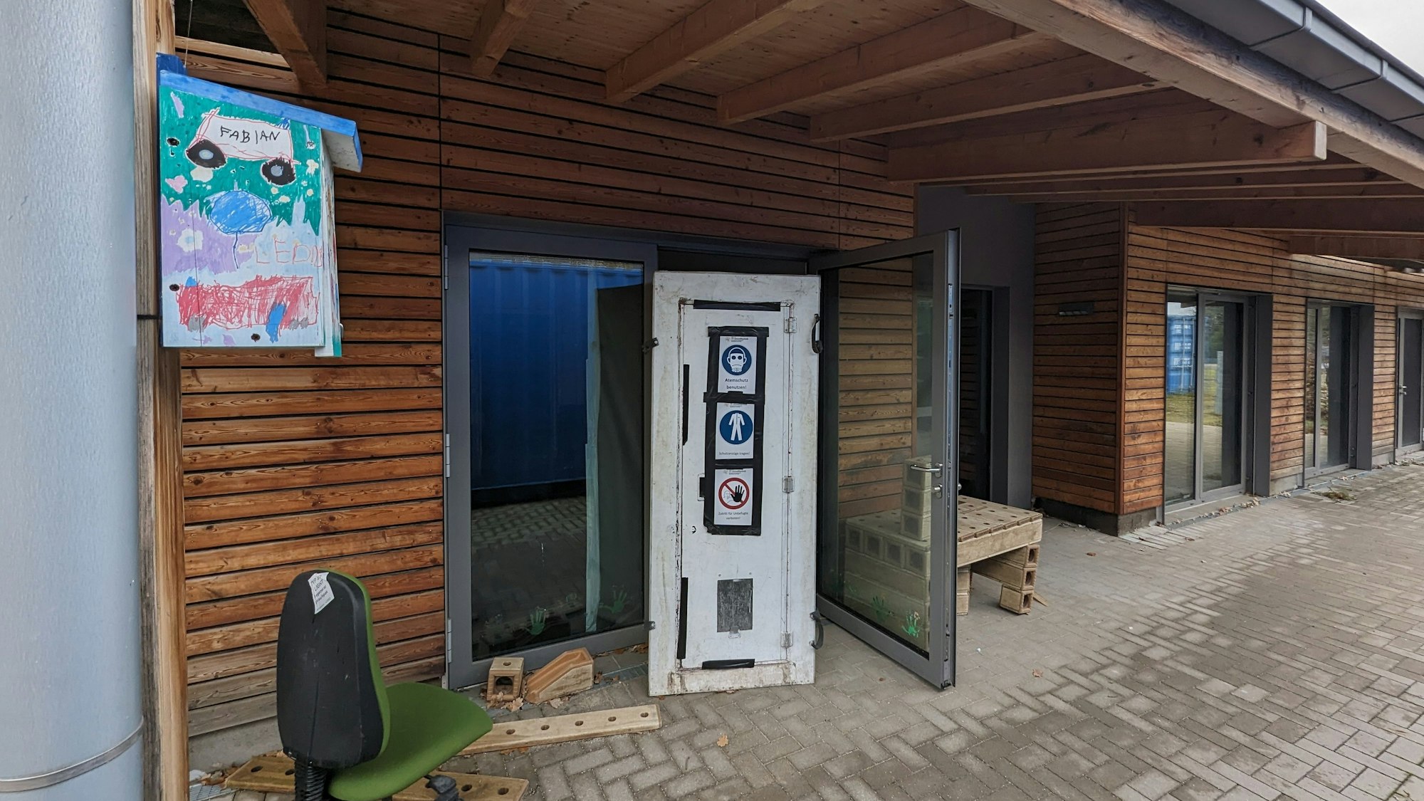 Eine Sicherheitsschleuse und ein Drehstuhl vor einem eingeschossigen Haus mit Holzfassade.