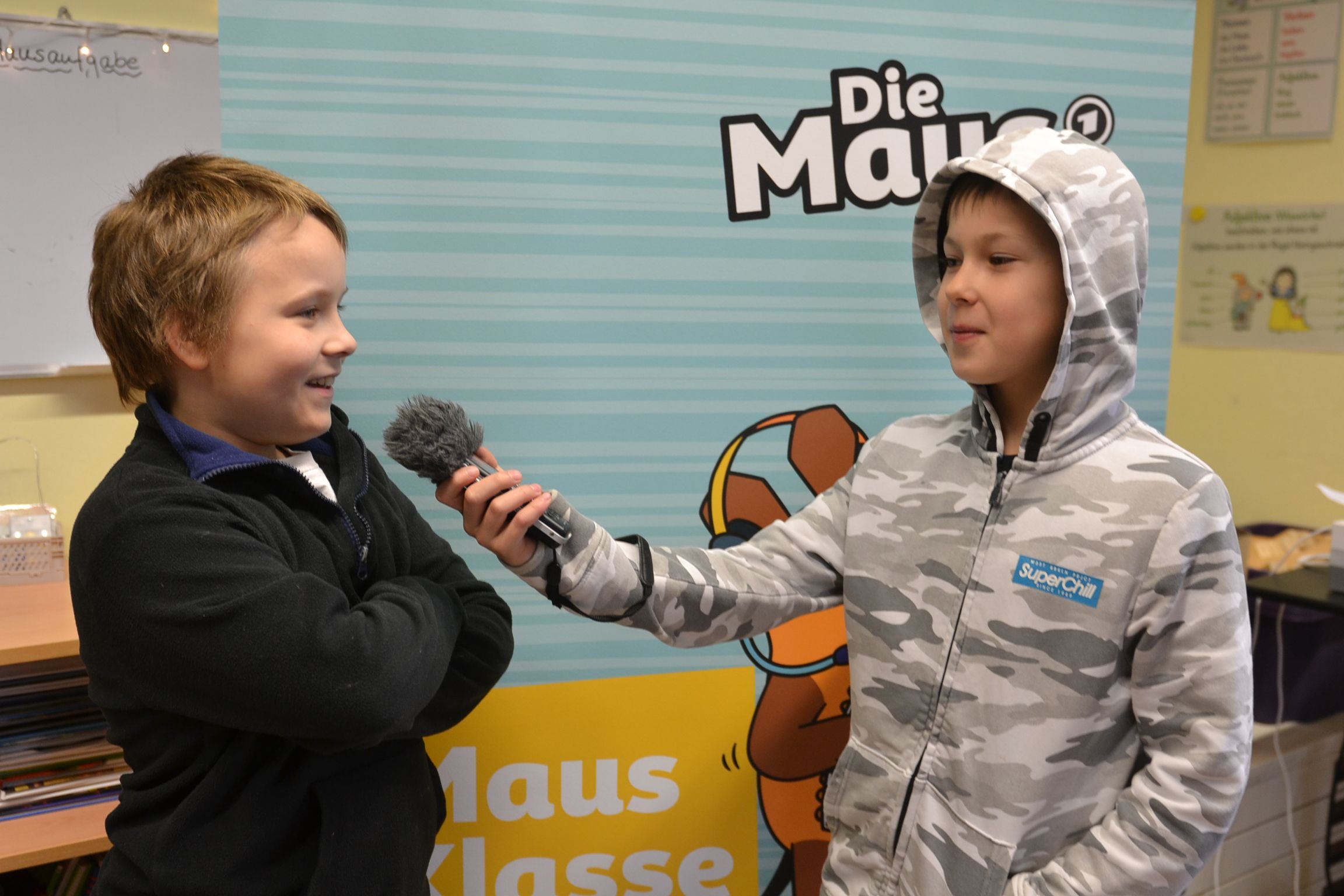 Zwei Schüler stehen vor einem Plakat der Sendung mit der Maus. Einer hält dem anderen ein Mikrofon hin.