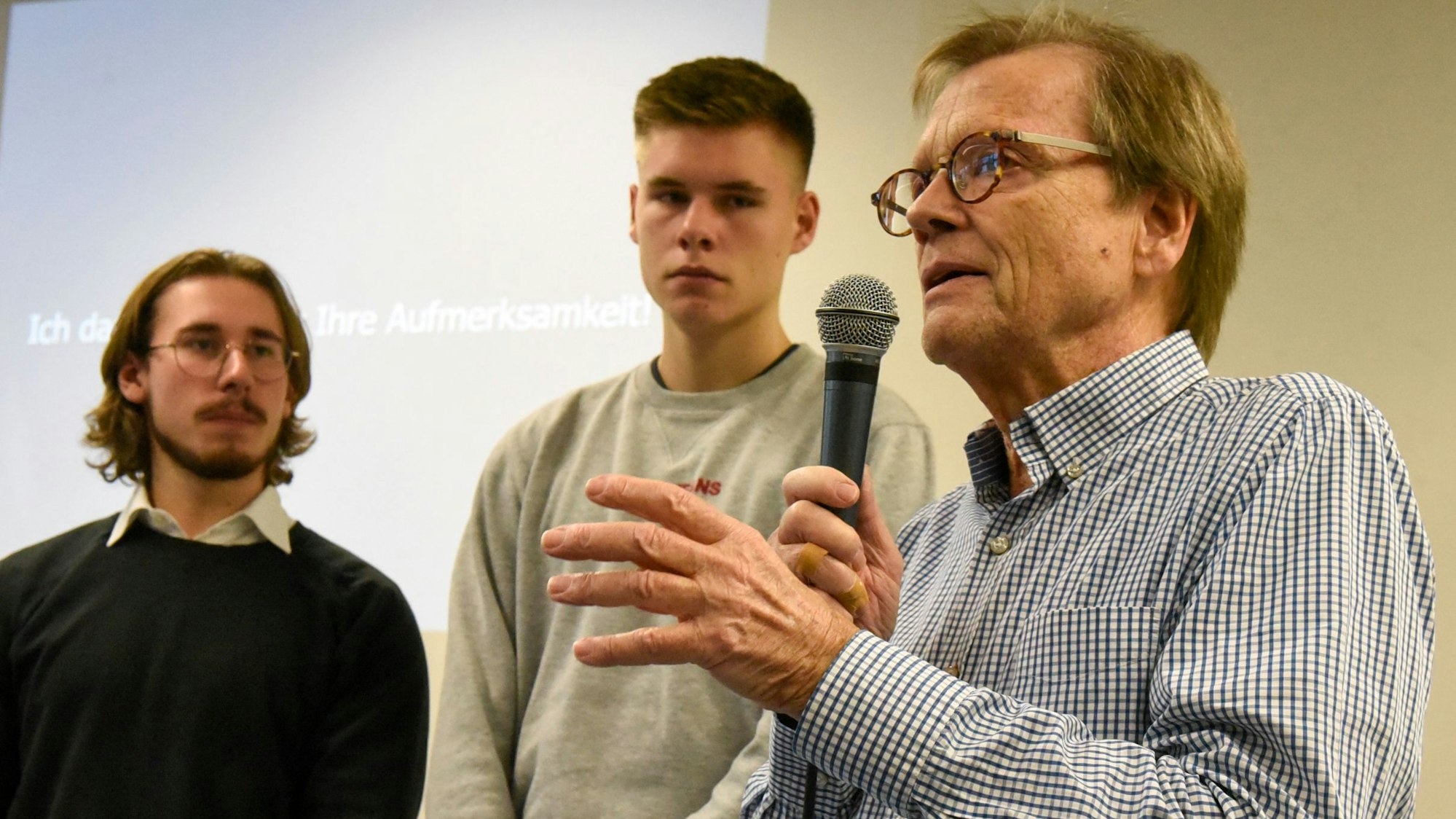Ein Mann am Mikrofon redet, junge Leute hören zu.