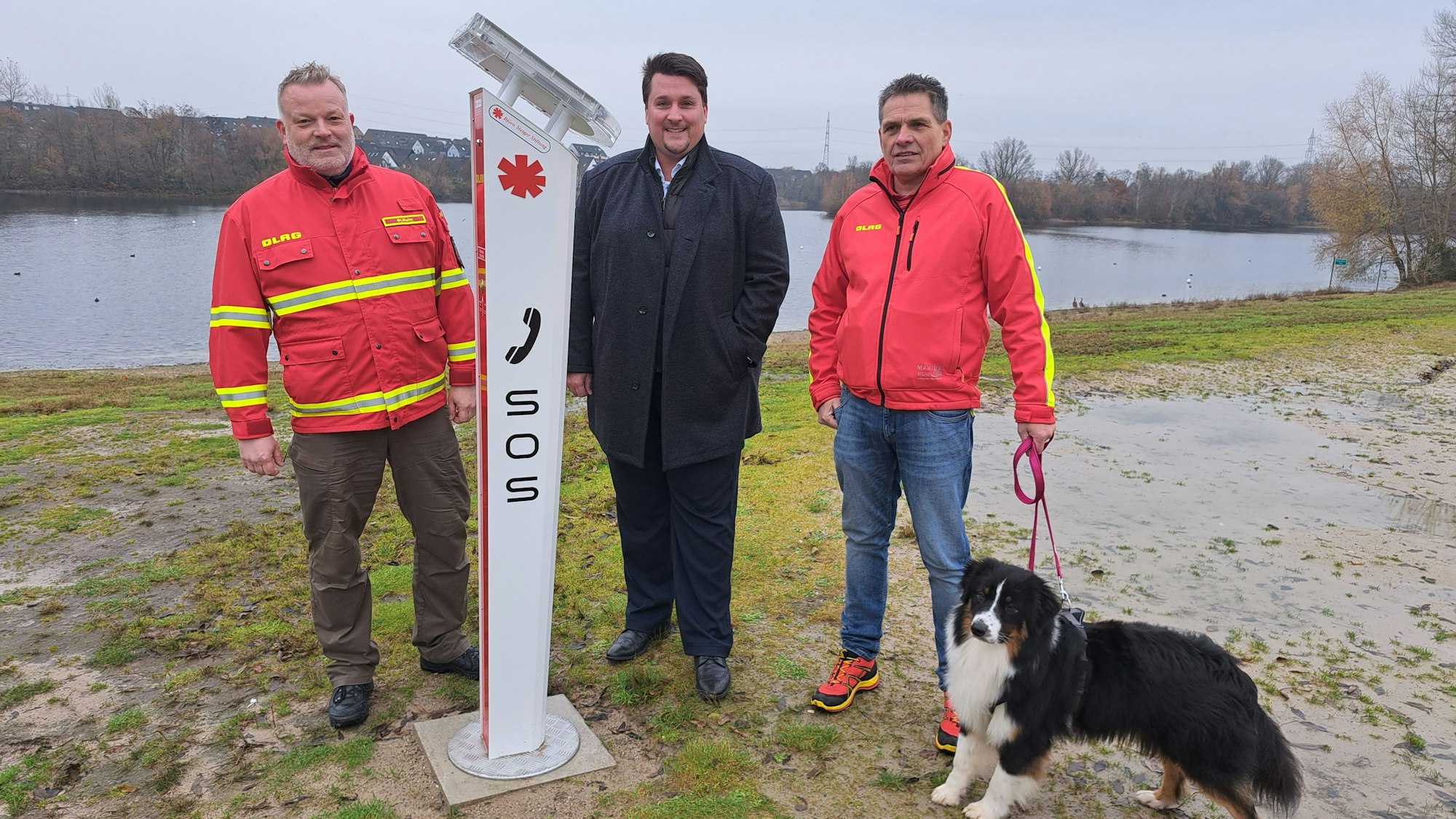 Stephan Halm, Pressesprecher der DLRG Rhein-Sieg, Troisdorfs Bürgermeister Alexander Biber und DLRG-Bezirksleiter Bruno Schöneberg stehen am Badestrand des Rotter Sees neben der neuen Notrufsäule, die dort mit Unterstützung der Björn-Steiger-Stiftung aufgestellt wurde.