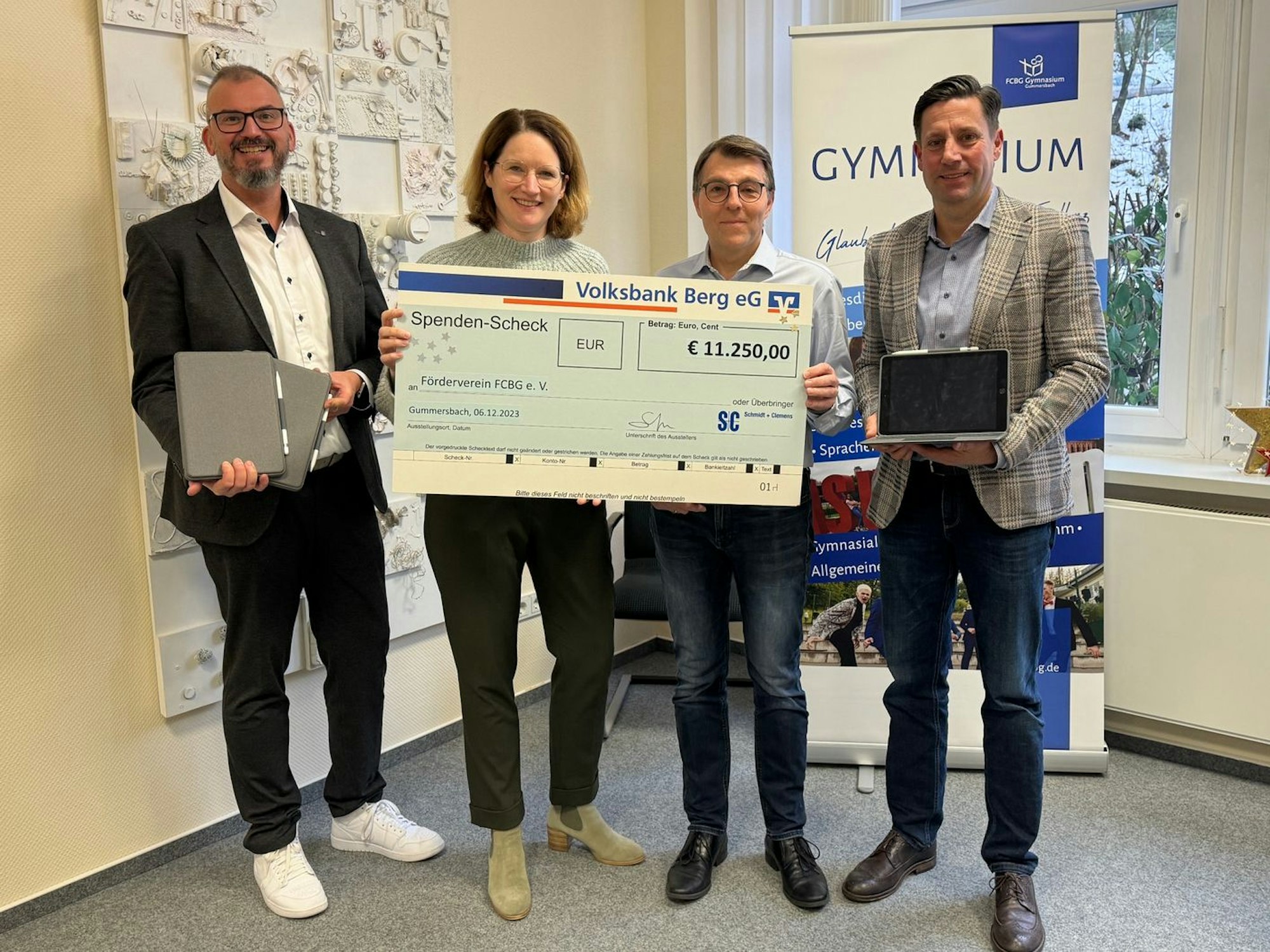 Spendenübergabe an die FCBG: Vier Personen sind auf dem Bild zu sehen, sie halten einen großen Spendenscheck in Händen, zwei zeigen iPads in die Kamera.