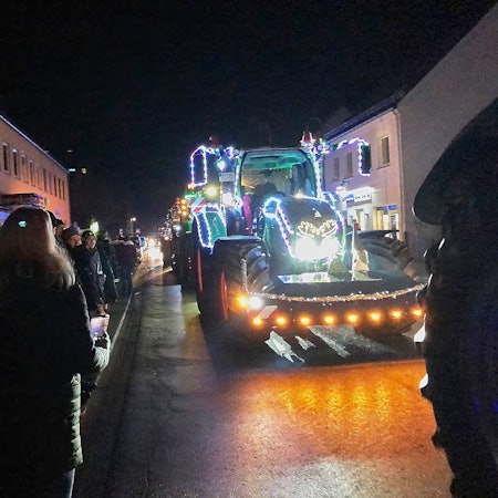 Ein mit Lichterketten behangener Traktor fährt über eine Straße, links und rechts stehen Schaulustige.