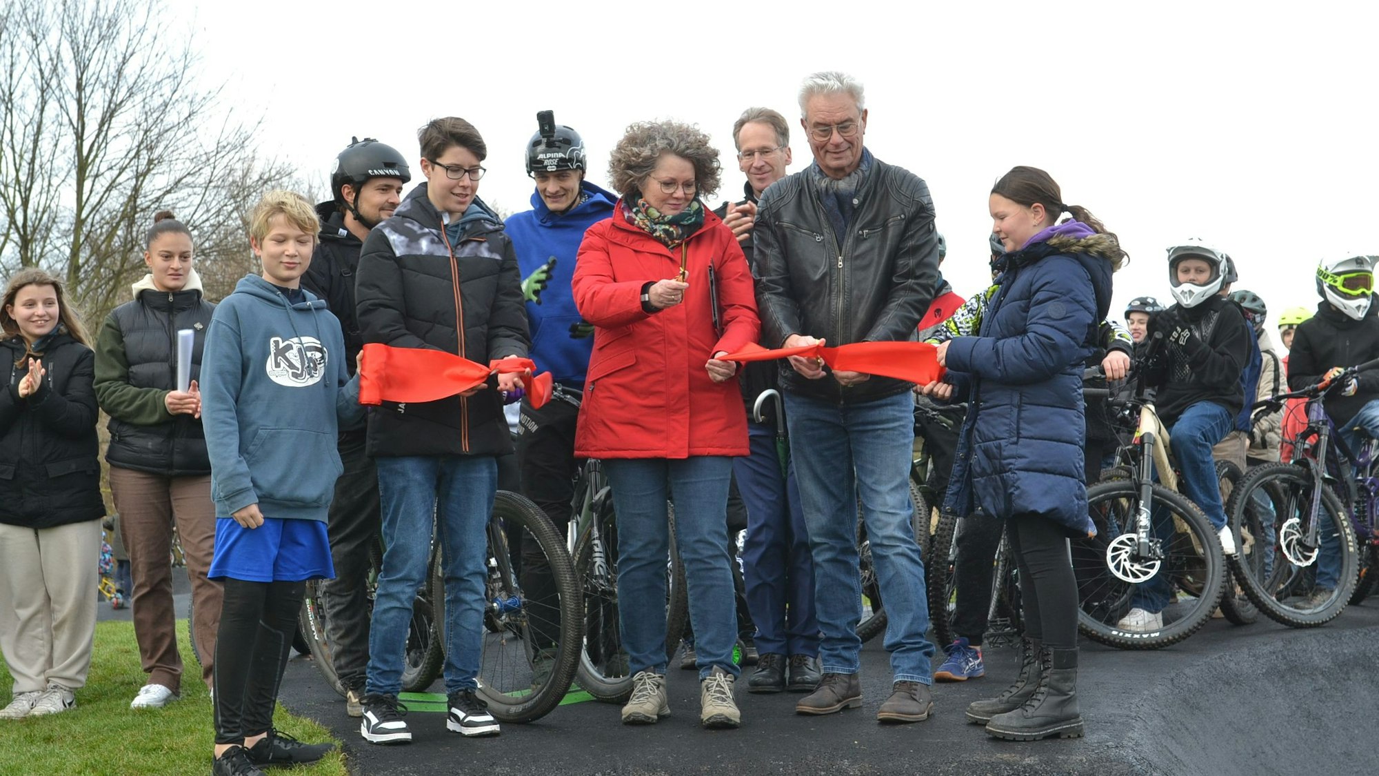 Bürgermeisterin Anne Horst, KJP-Vorsitzender Simon Krämer und der stellvertretende Vorsitzende der LEADER-Region Albert Bergmann stehen auf einem Stück Asphalt und haben ein rotes Band in der Hand. Um sie herum stehen viele Kinder und Jugendliche mit Fahrrädern.