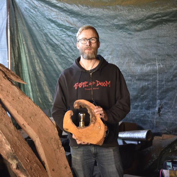 Björn Schütz steht in einem Schuppen neben alten Holzbalken. In der Hand hält er eine Lampe aus einer Baumscheibe.