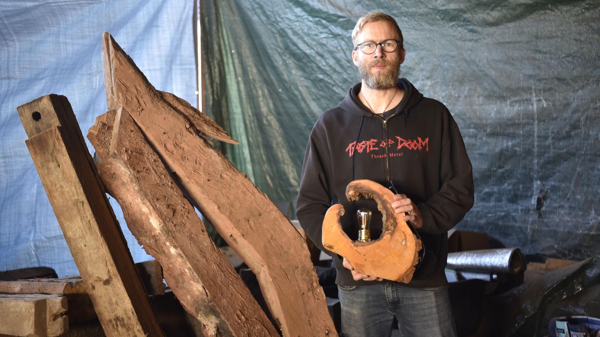 Björn Schütz steht in einem Schuppen neben alten Holzbalken. In der Hand hält er eine Lampe aus einer Baumscheibe.