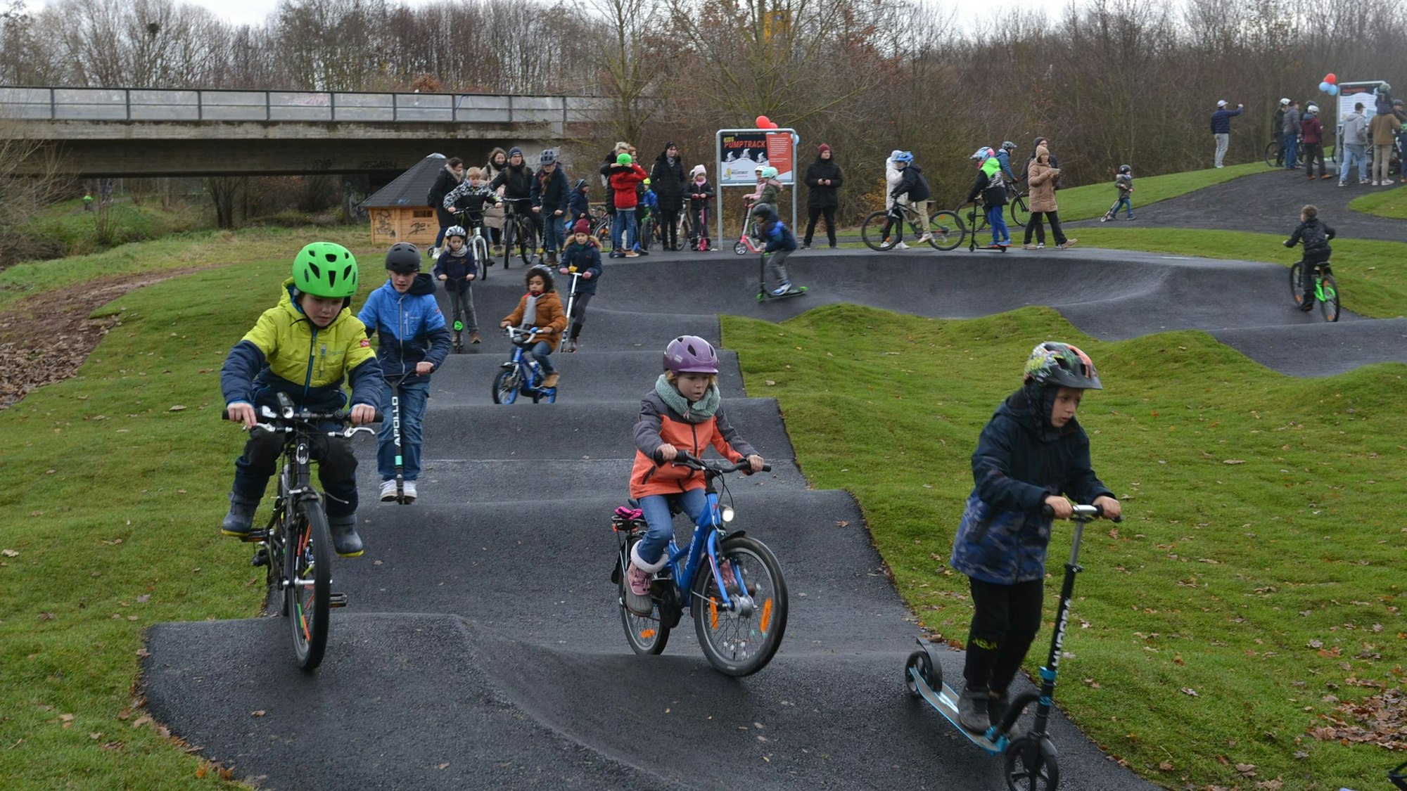 Kinder fahren mit Fahrrädern und Rollern über eine Pumptrack-Anlage.
