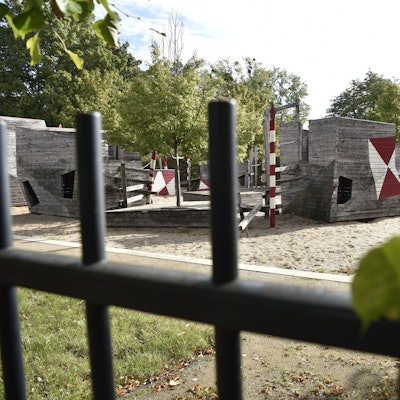 Hinter einem Zaun sind Spielgeräte aus Holz zu erkennen.