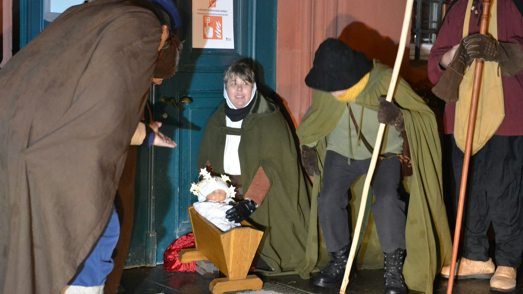 Mehrere Menschen in Kostümen stehen um eine Puppenwiege herum, in der eine Puppe als Jesuskind liegt.