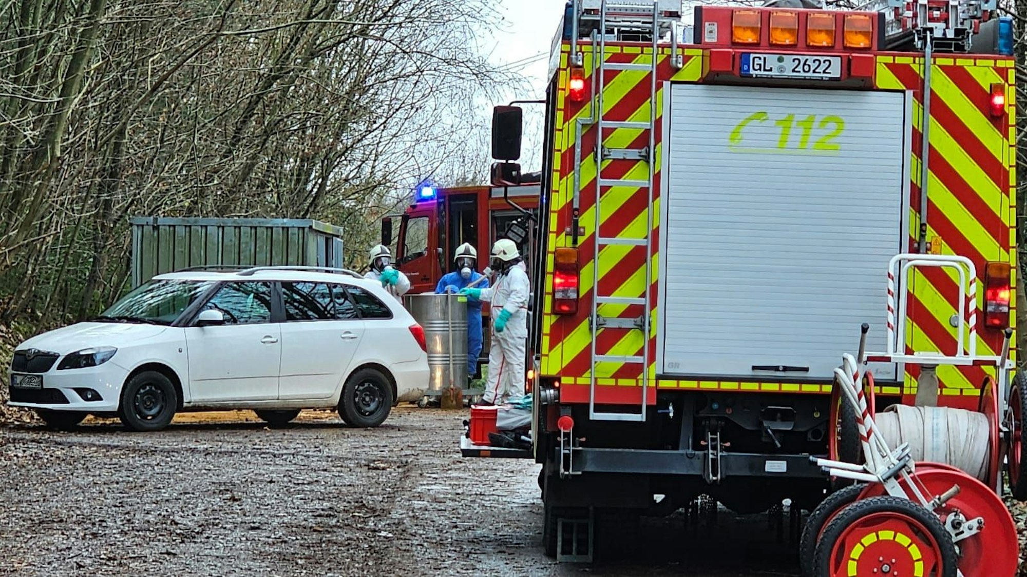 Feuerwehrleute in Schutzanzügen und mit Atemschutz stehen vor einem Feuerwehrfahrzeug an einem Metallfass-