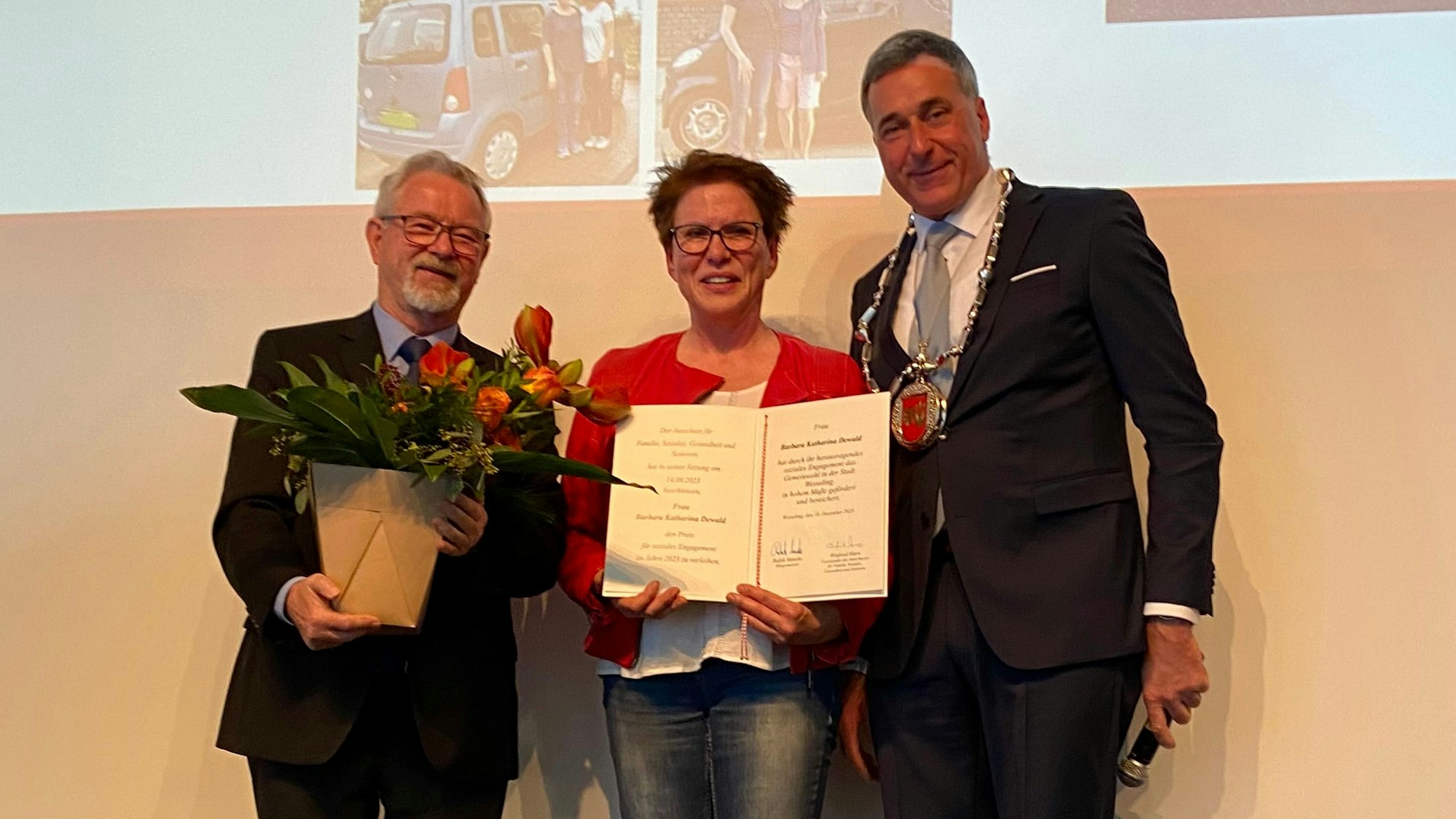 Zwei Männer überreichen einer Frau eine Urkunde und Blumen.