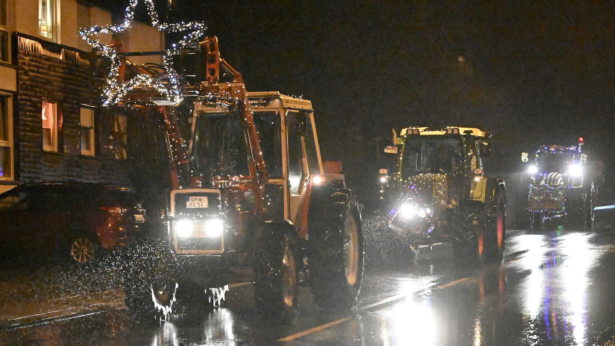Mit Lichterketten geschmückte Traktoren fahren über eine verregnete Landstraße.