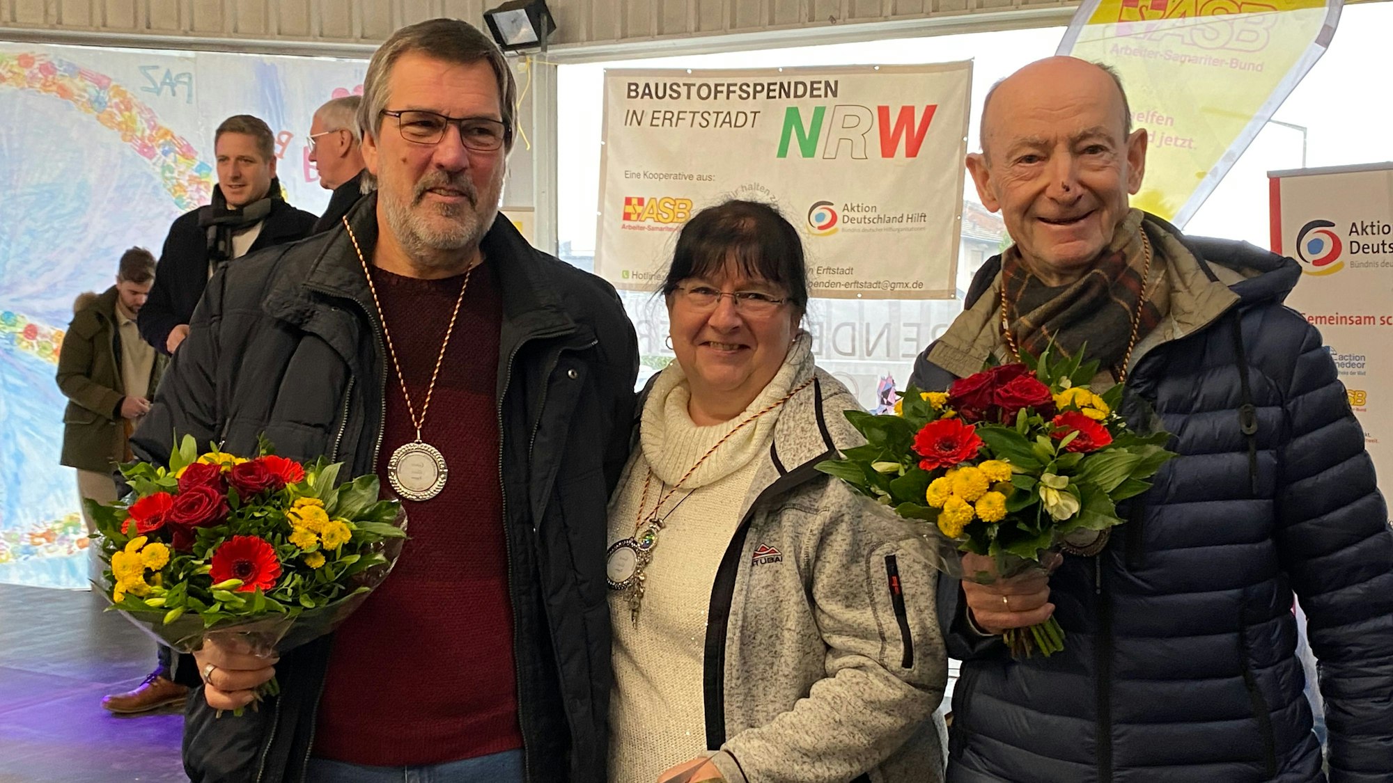 Eine Frau und zwei Männer halten Blumensträuße in den Händen.
