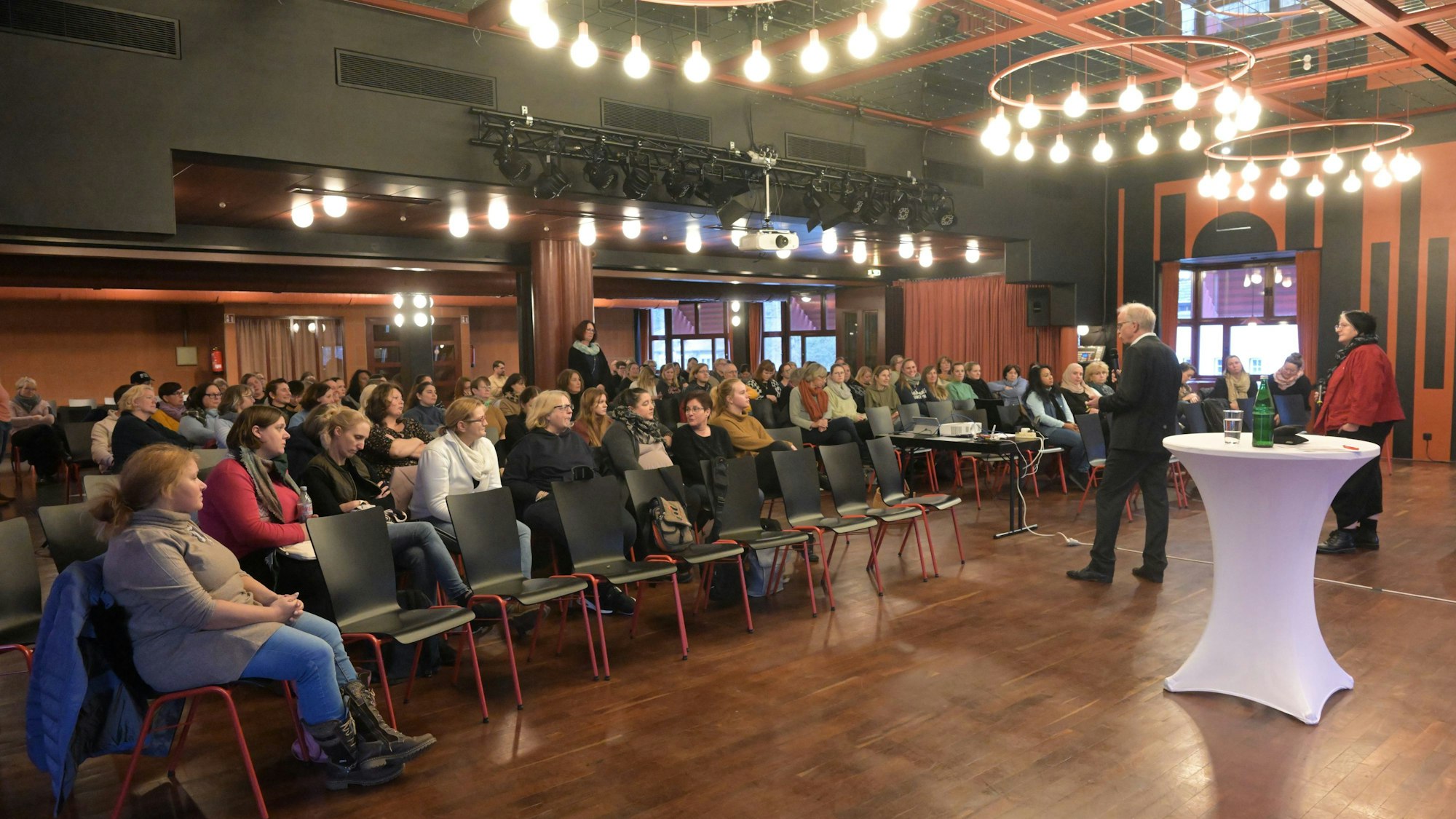Ein Redner und eine Rednerin stehen vor den besetzten Stuhlreihen.