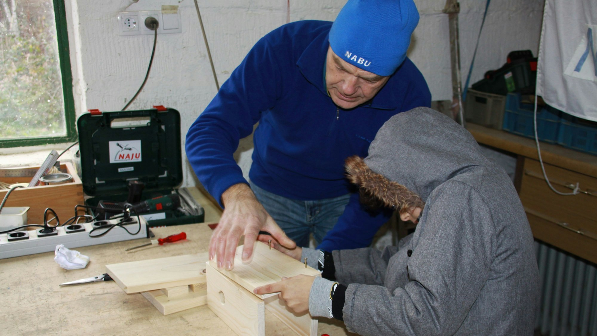 An einer Werkbank hilft ein Mann vom Nabu einem Kind beim Zusammensetzen eines Nistkastens aus Holz.