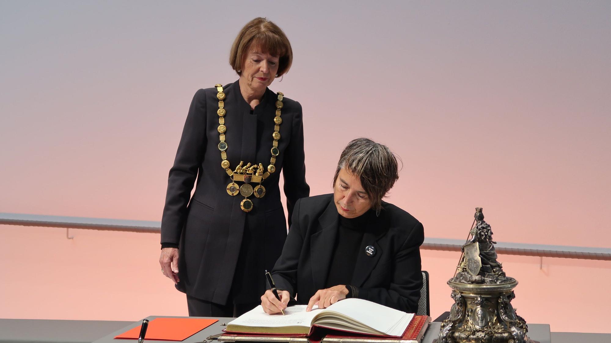 10.12.2023 Dr. Monika Hauser trägt sich in das goldene Buch der Stadt ein. Dabei OB Henriette Reker.