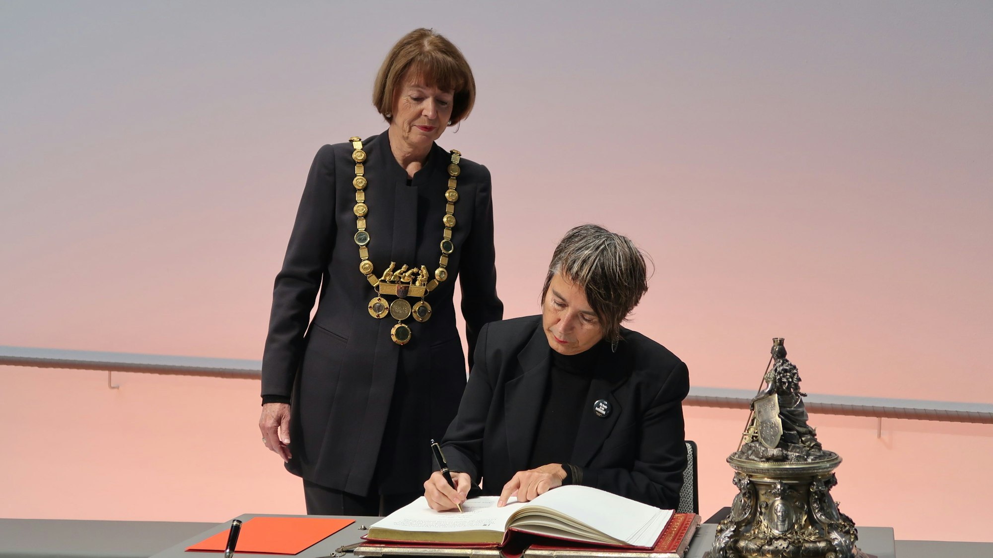 10.12.2023 Dr. Monika Hauser trägt sich in das goldene Buch der Stadt ein. Dabei OB Henriette Reker.